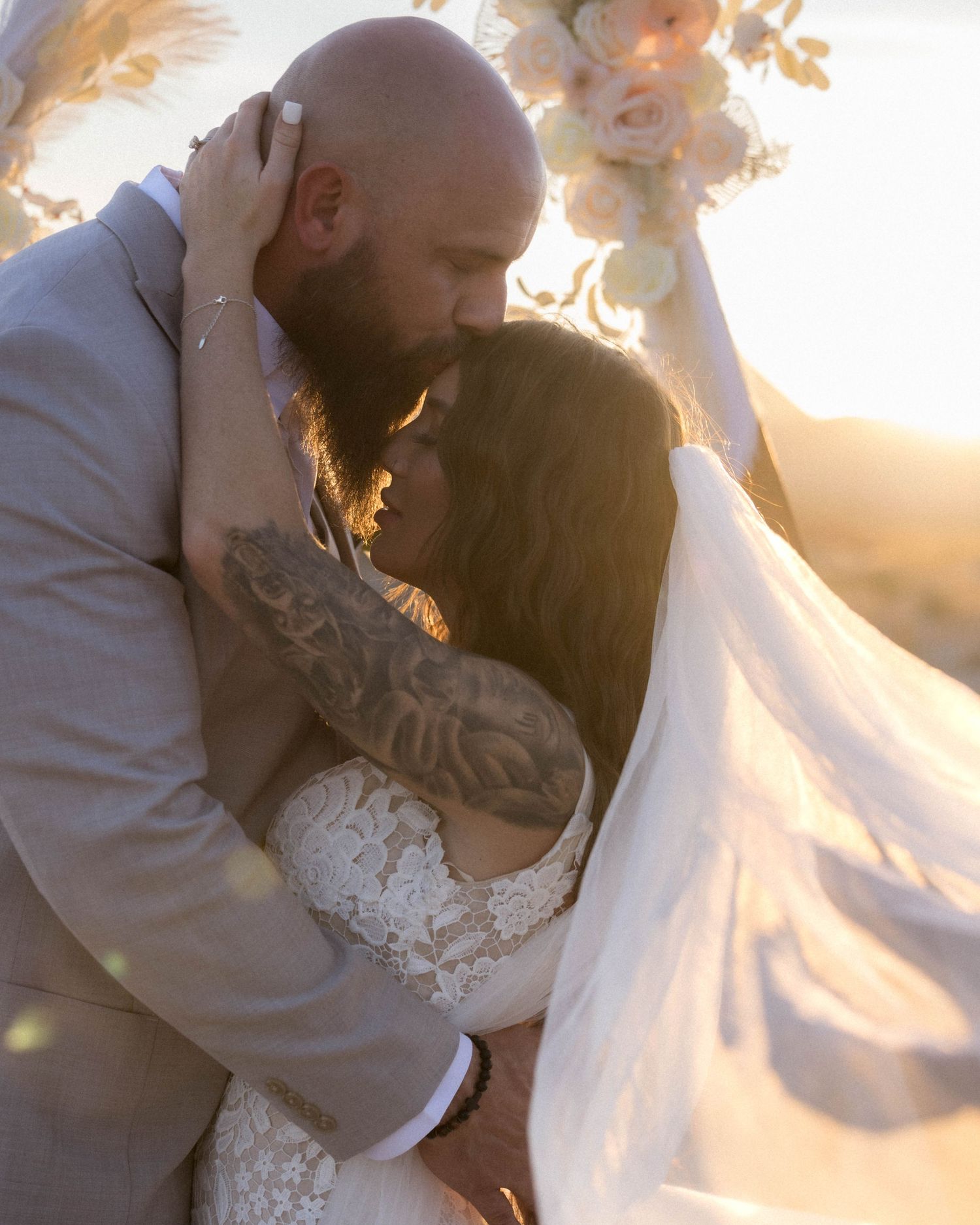 Tattooed bride and bearded groom share a romantic kiss under floral arch at golden sunset wedding.