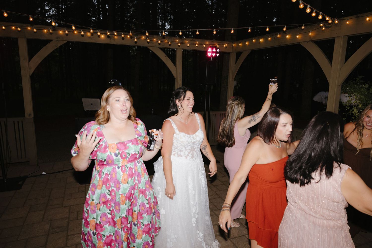 Bride in white lace gown dances joyfully with guests under string lights at an outdoor evening wedding reception.