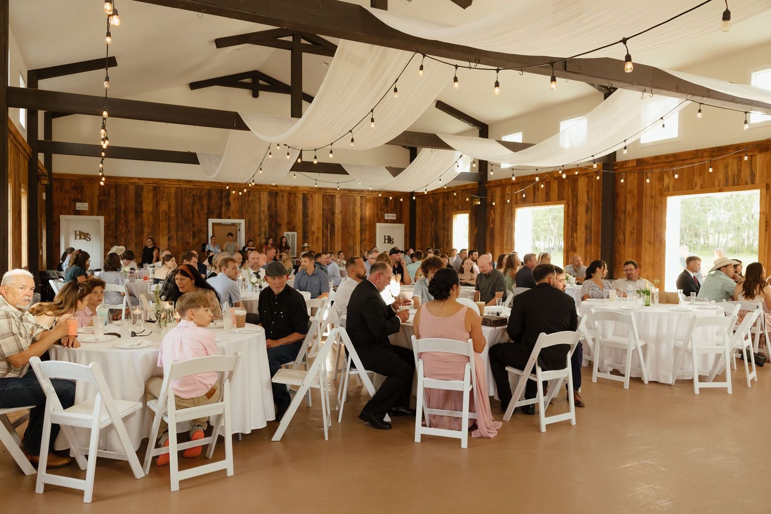Guests dine at round tables in rustic barn wedding reception with string lights above.