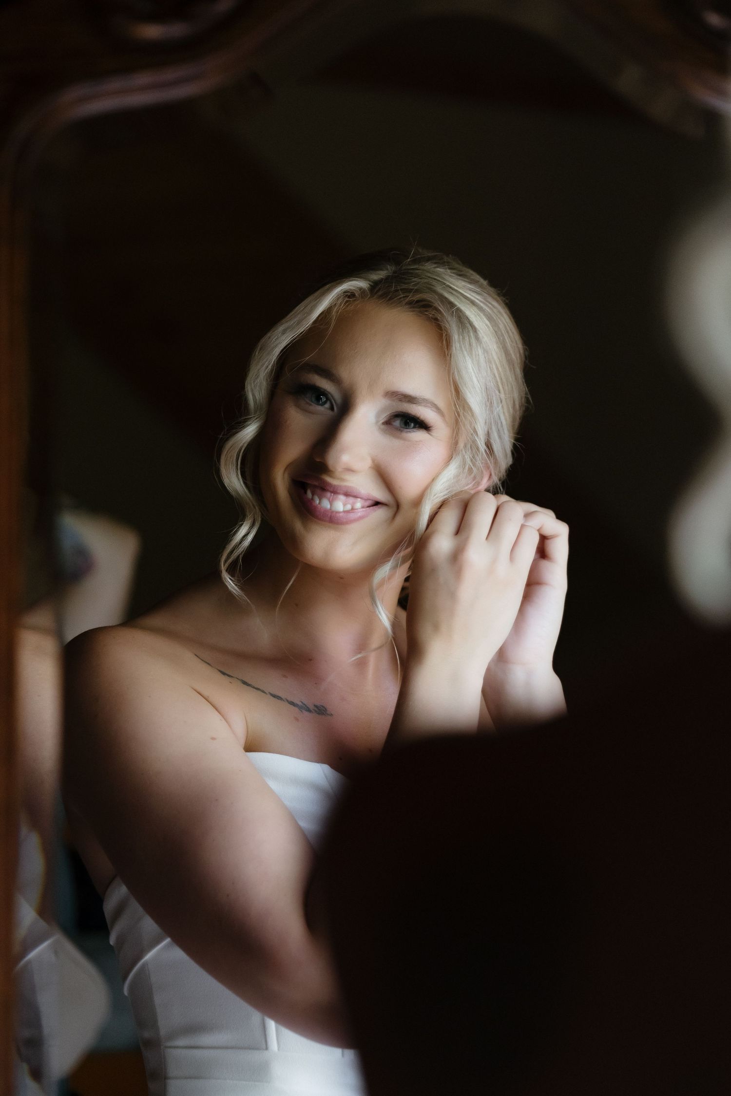 Smiling bride putting on earrings while getting ready, reflected in a vintage mirror on her wedding day.