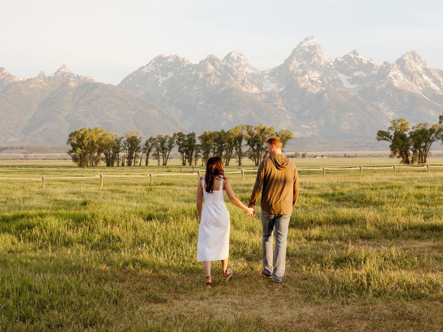 Couple holding hands walking through a sunlit meadow with the Grand Teton mountains in the distance.