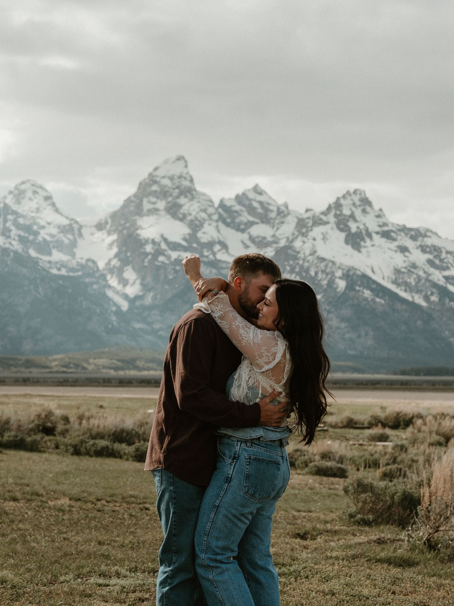 Couple sharing a romantic kiss in a field with snow-capped Grand Teton mountains in the background.