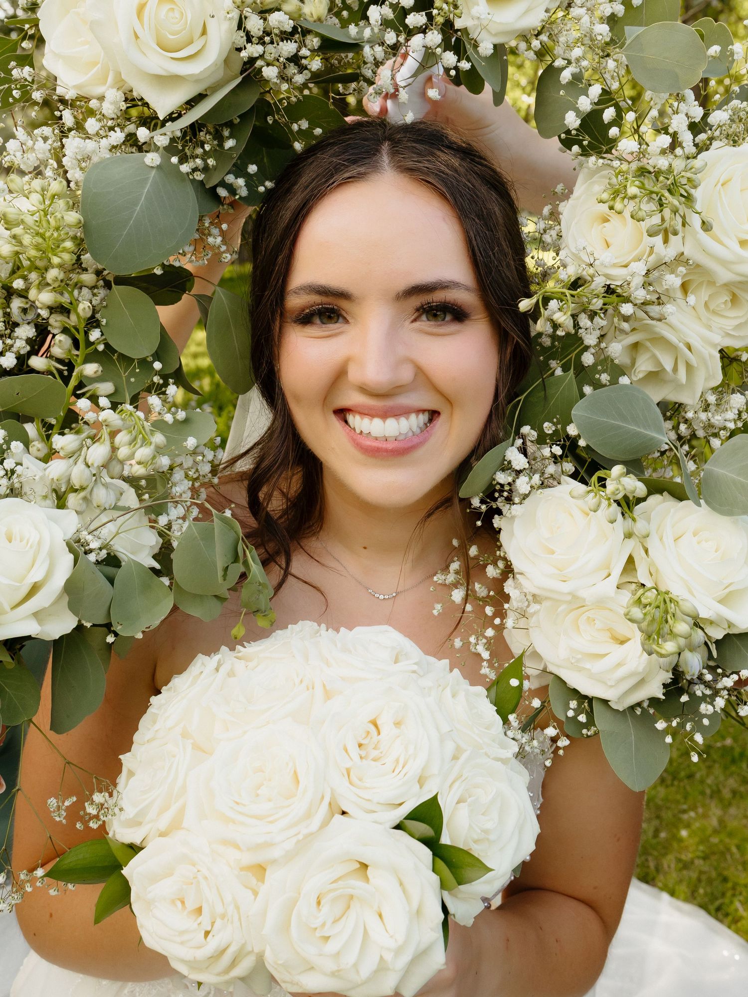 Smiling bride surrounded by white roses, baby's breath, and eucalyptus bouquets on her wedding day.
