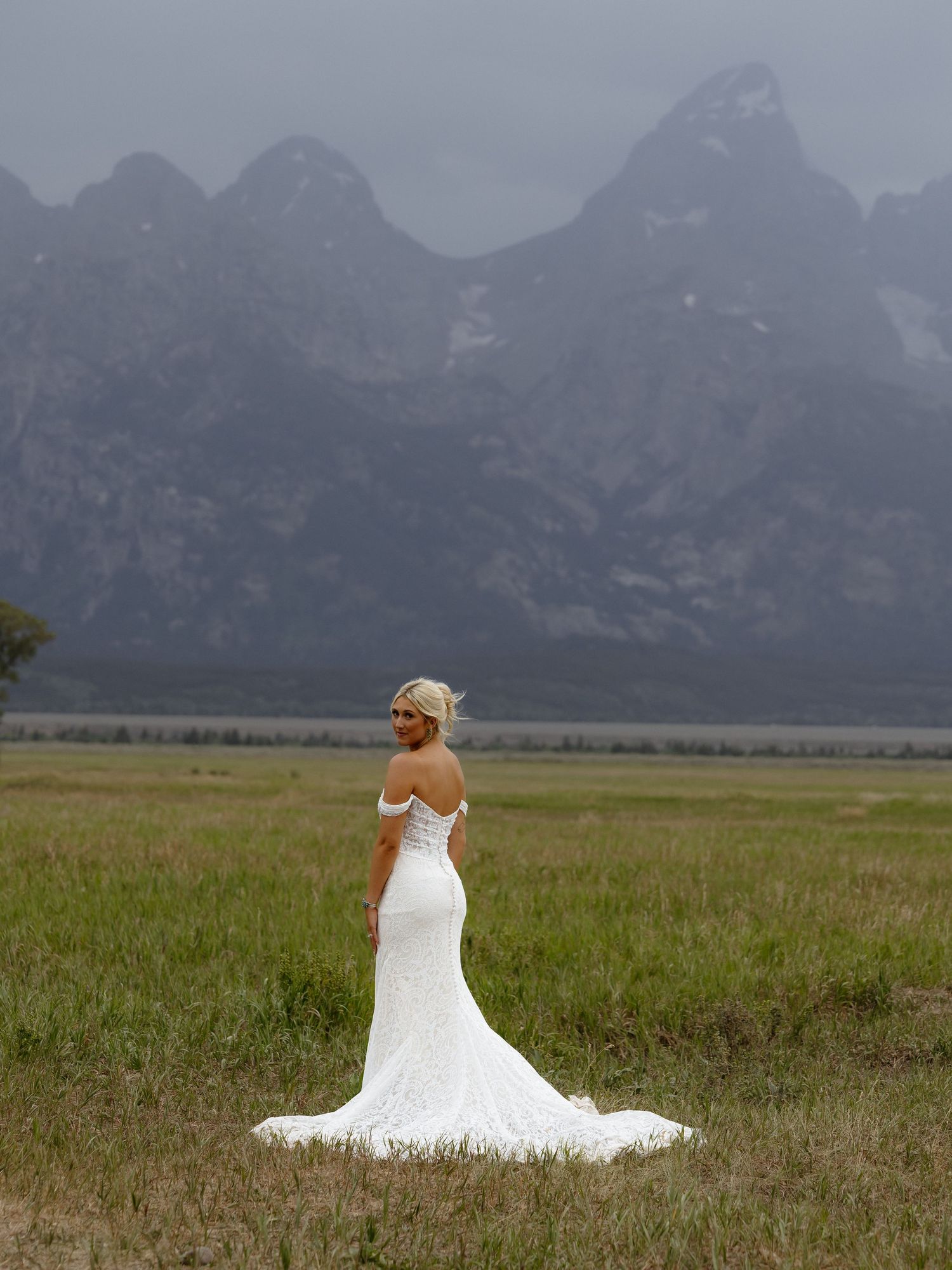 Bride in white off-shoulder gown stands in meadow with dramatic mountain backdrop on overcast day.