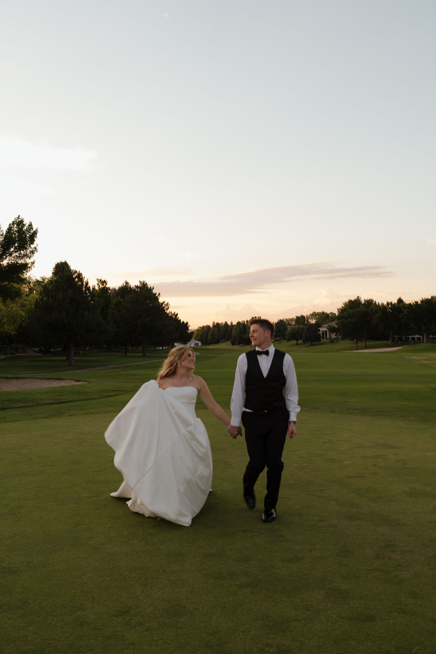 Bride and groom walk hand-in-hand across a lush green golf course at sunset, bride in white gown, groom in black tuxedo.