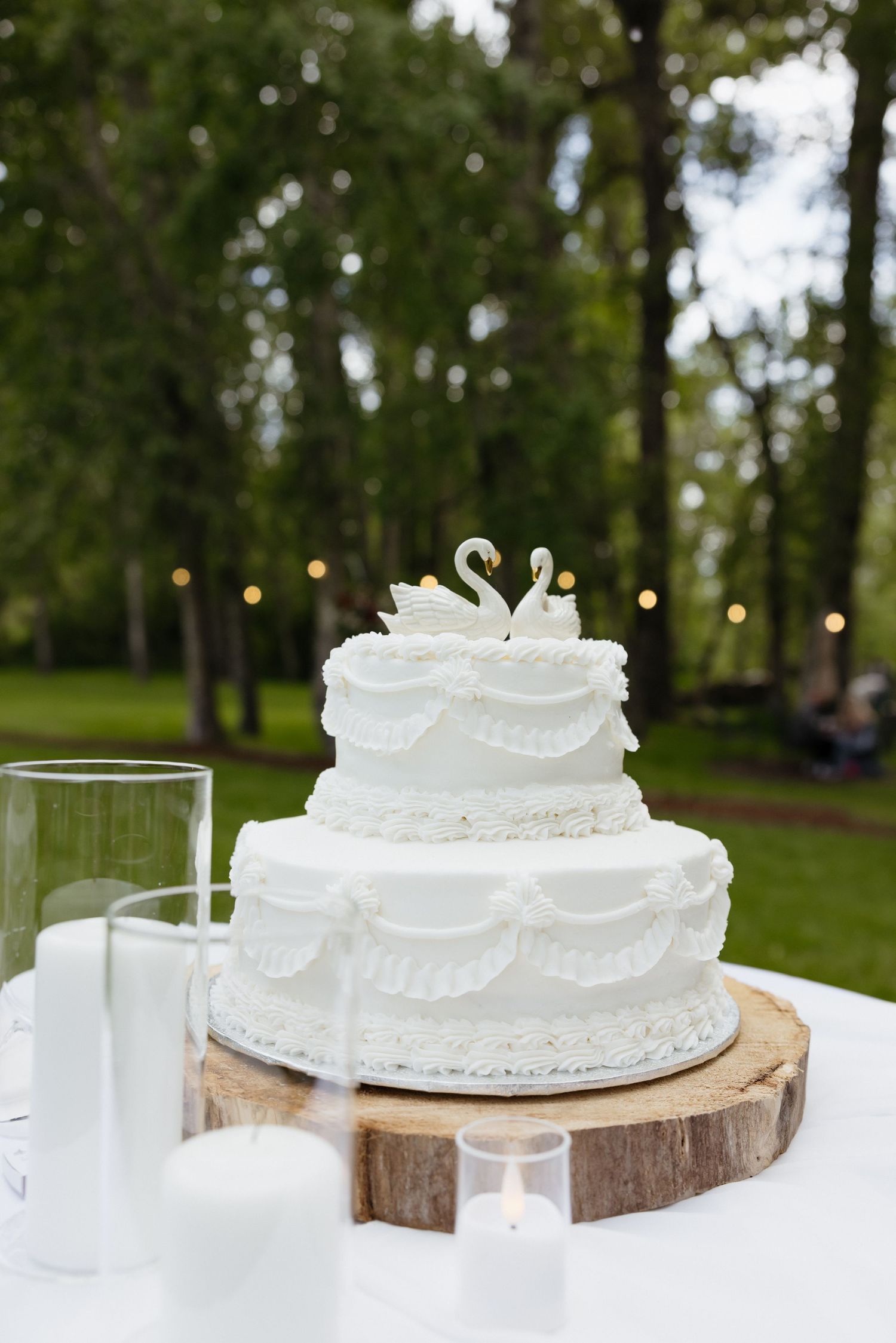 Classic two-tier white wedding cake with swan cake toppers on wooden slice stand at outdoor garden reception.