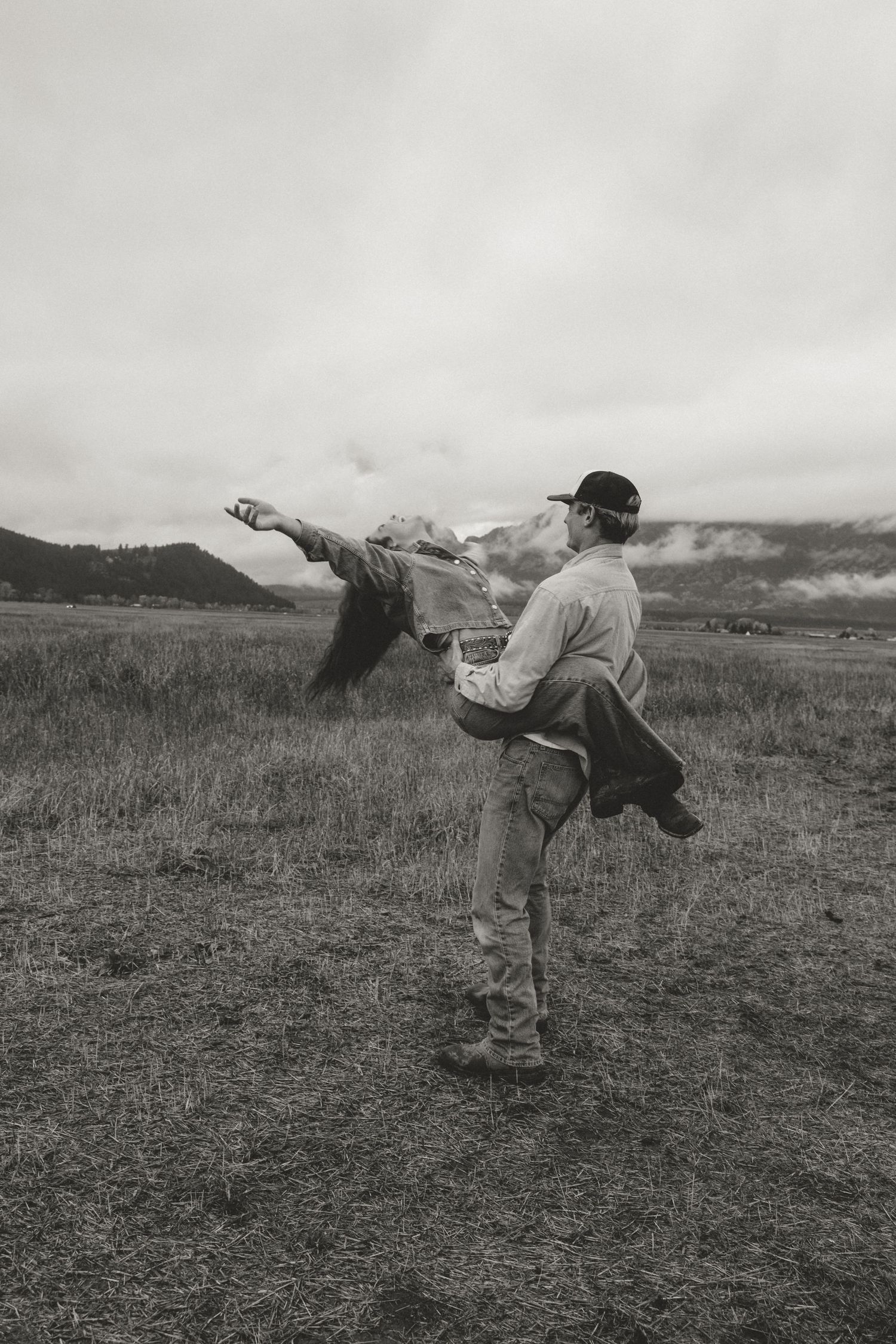 Black and white photo of man carrying woman in a field with mountain landscape behind them.