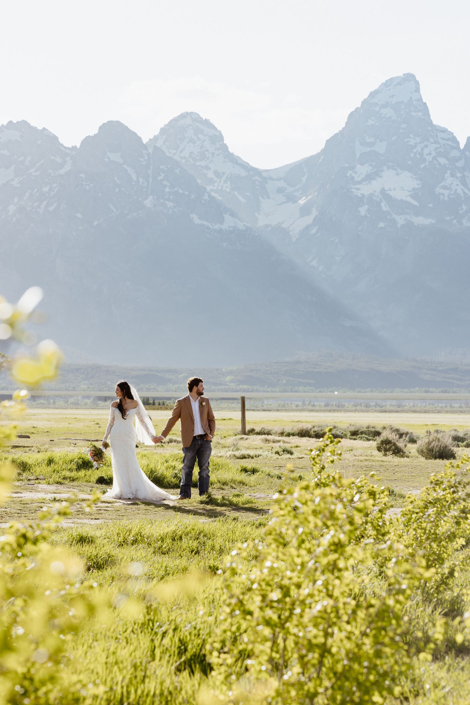 Bride and groom holding hands walking through meadow with snow-capped Grand Teton mountains behind.
