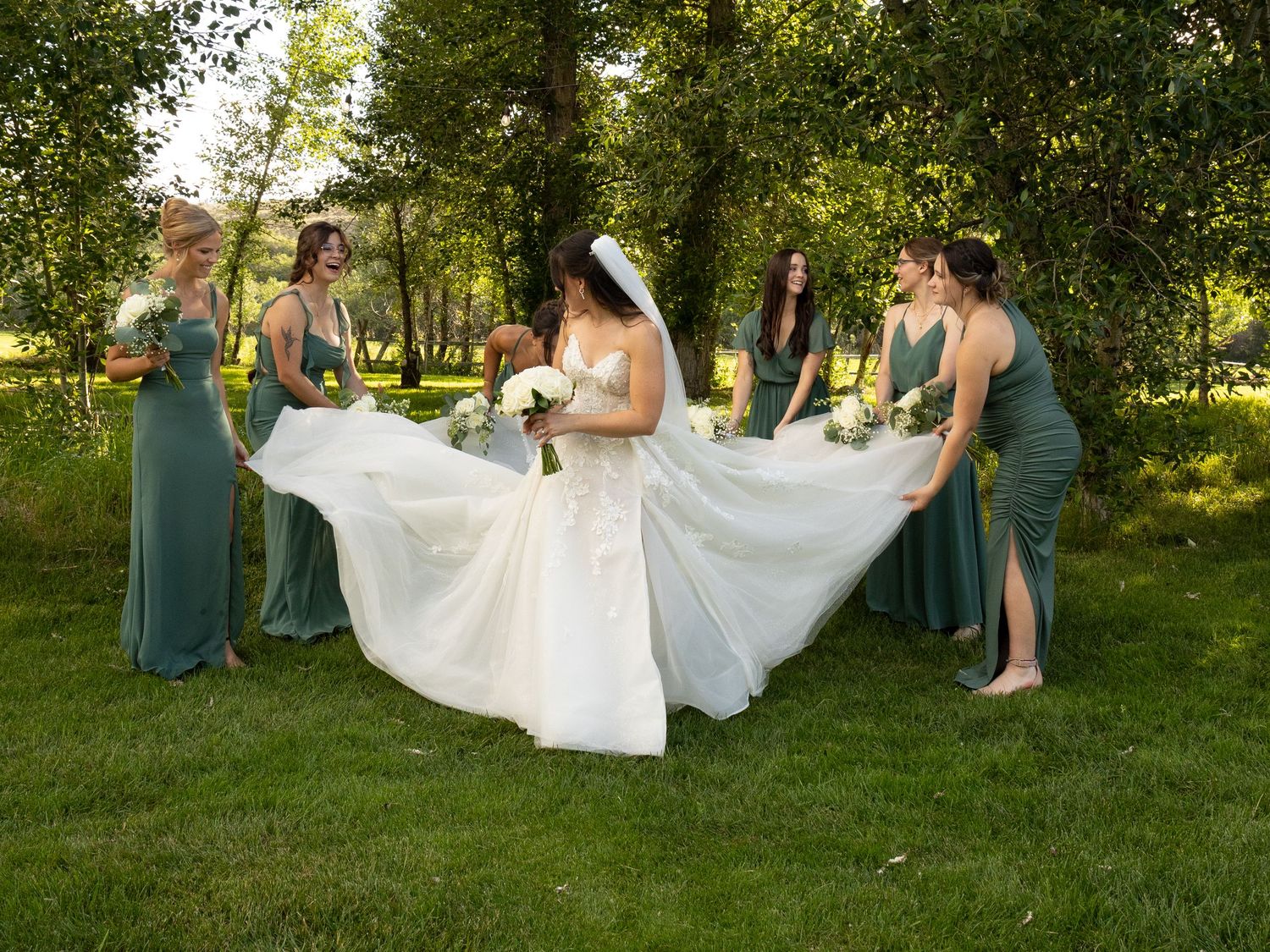 Bride in white gown with veil surrounded by bridesmaids in sage green dresses on lush green lawn.