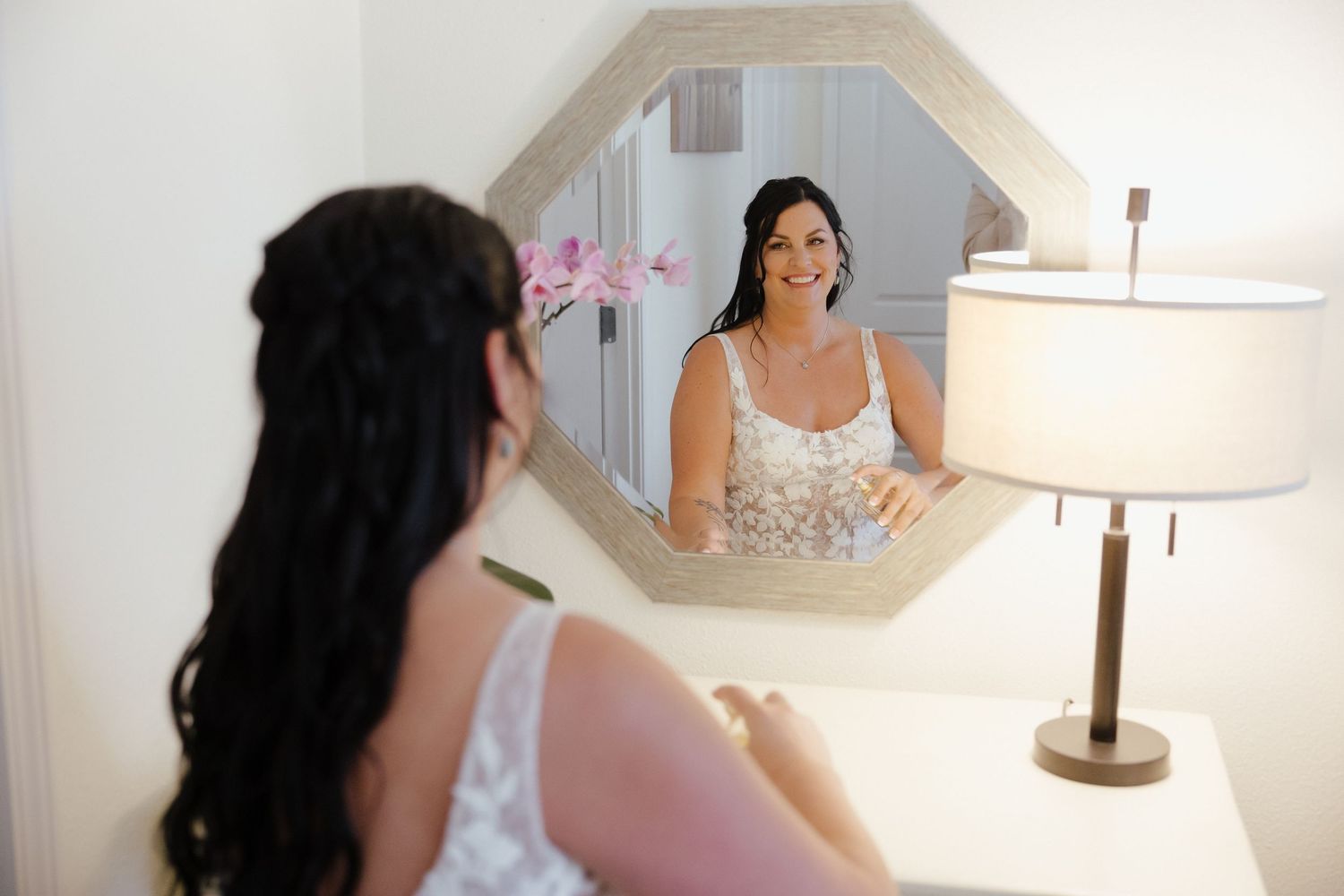 Bride in lace wedding dress smiling at her reflection in an octagonal mirror while getting ready.