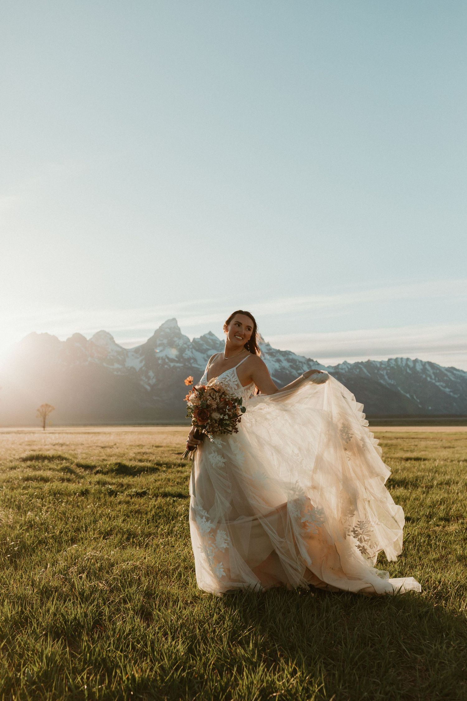 Bride in a flowing lace wedding gown holding a bouquet in a golden meadow with mountain peaks behind her.