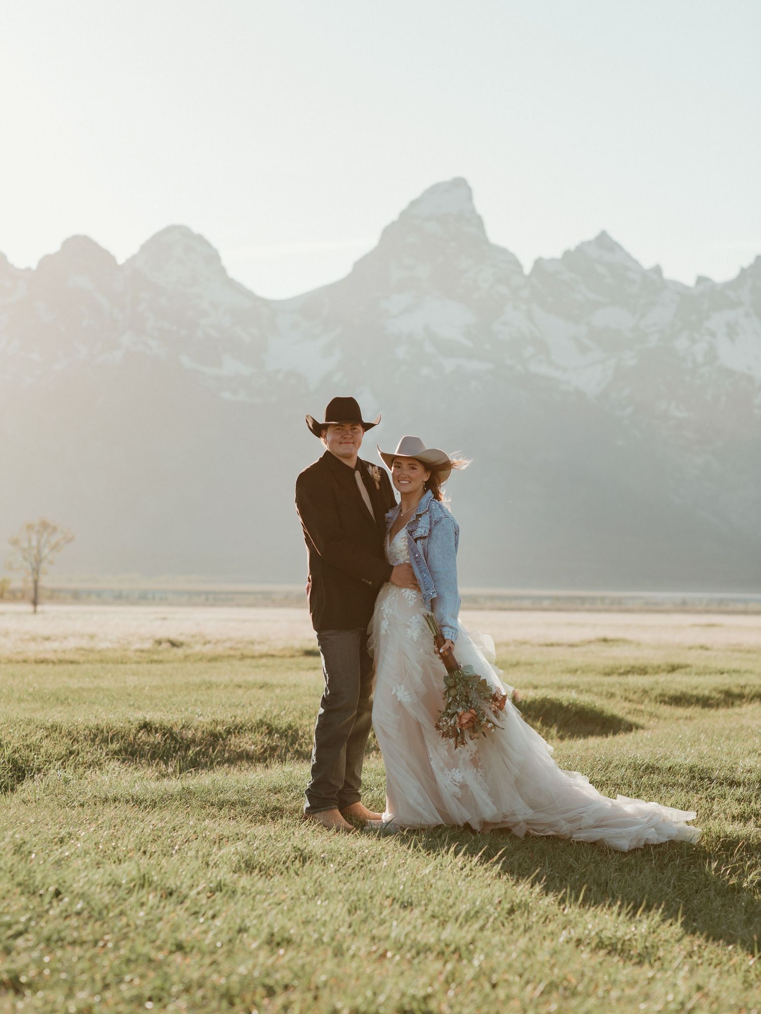 Couple in western attire with cowboy hats poses in Grand Teton meadow during golden hour wedding.
