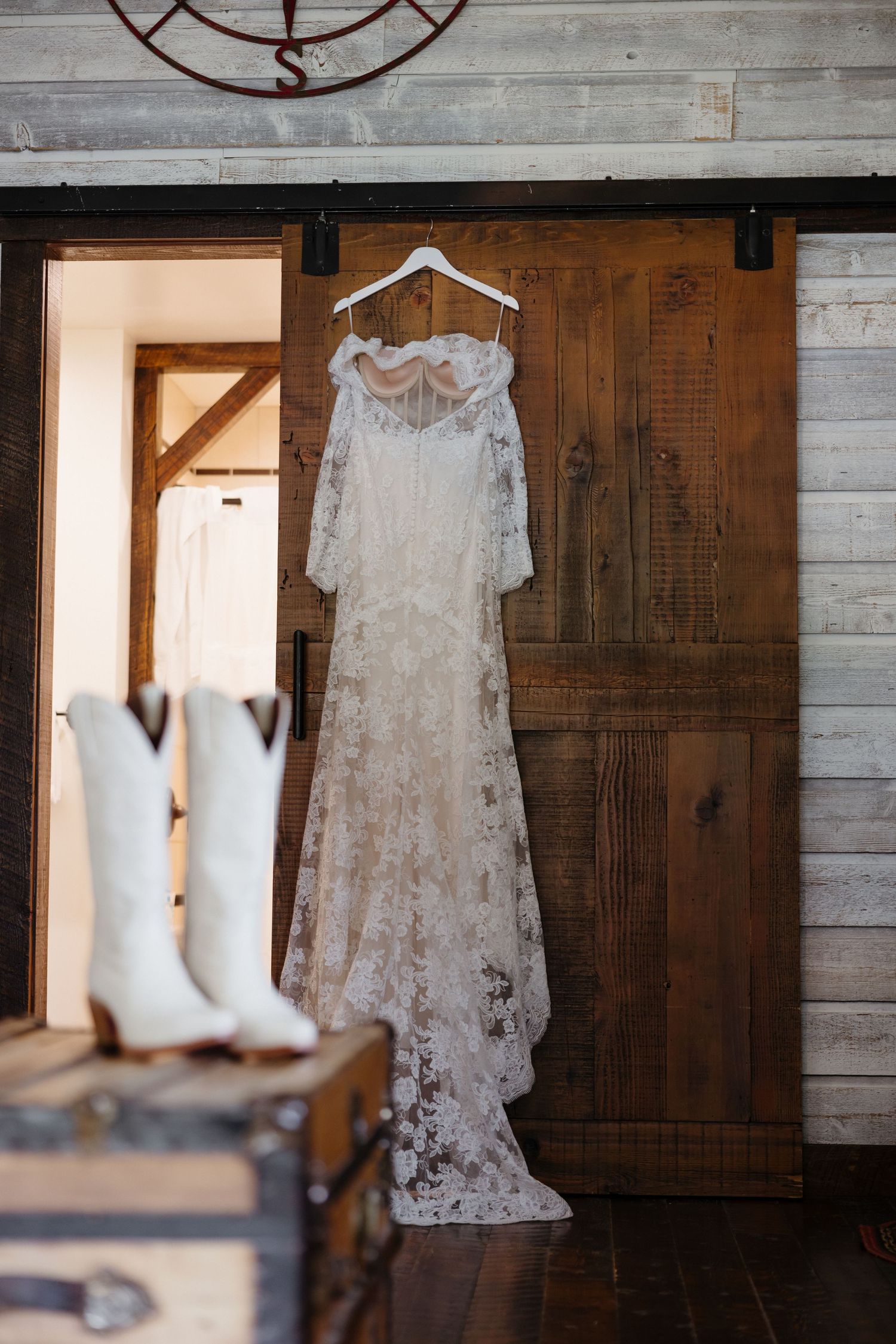 Lace wedding dress hanging on a barn door with white cowboy boots and vintage trunks in foreground.