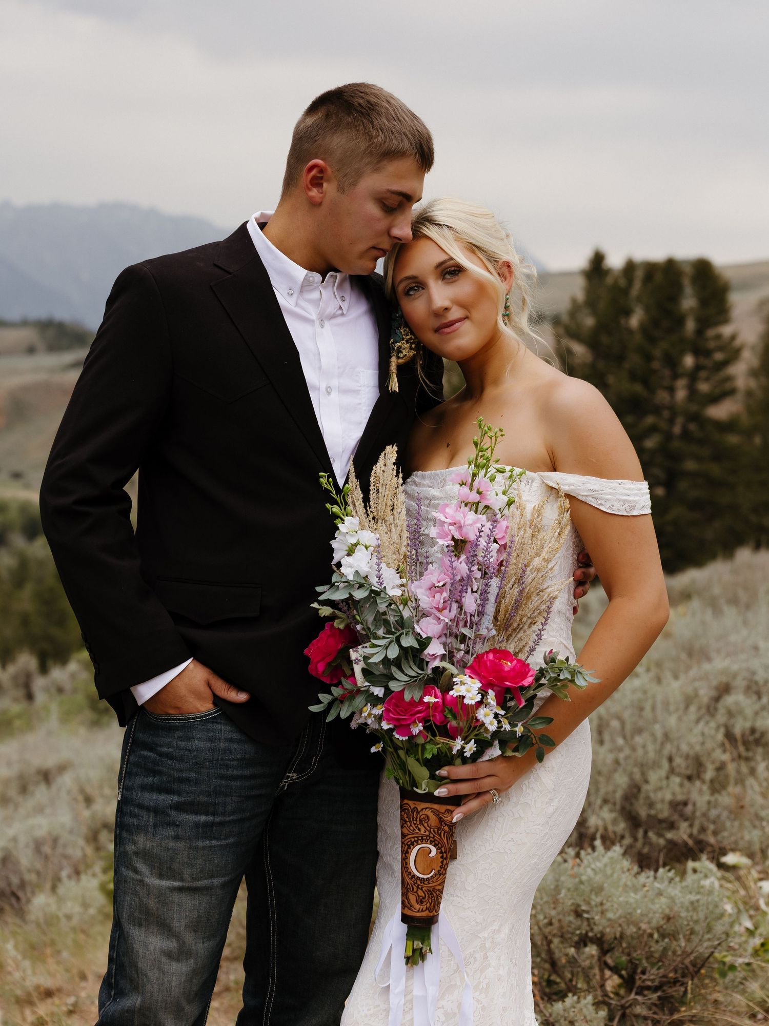 Bride and groom pose outdoors with mountain backdrop, she holds pink floral bouquet, he wears black suit with jeans.