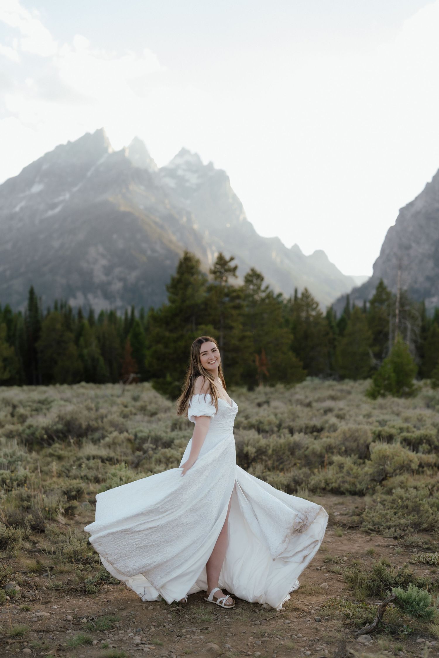 Bride in flowing white off-shoulder gown twirls in front of dramatic Grand Teton mountain landscape at dusk.