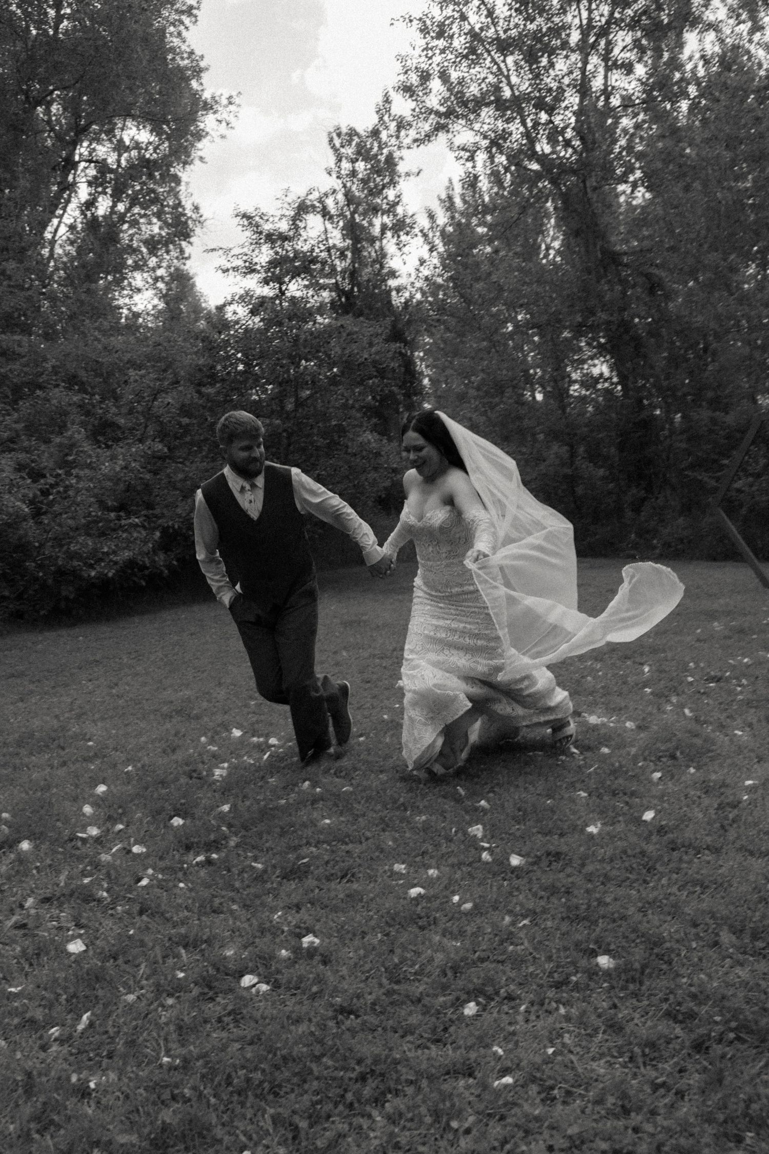 Black and white photo of bride and groom joyfully running hand in hand across a grassy outdoor setting.