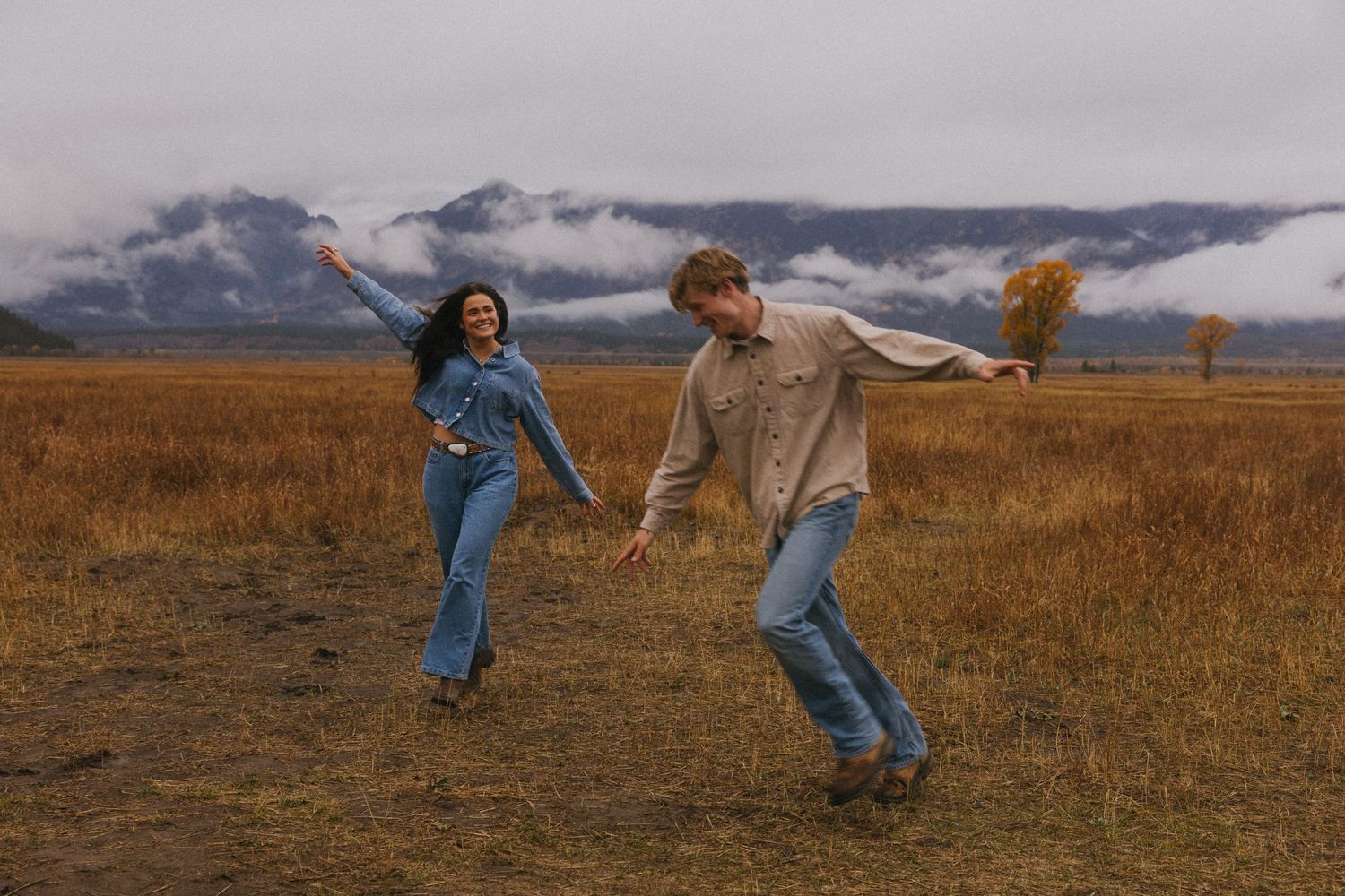 Playful couple holding hands and dancing in a golden autumn field with misty mountains.