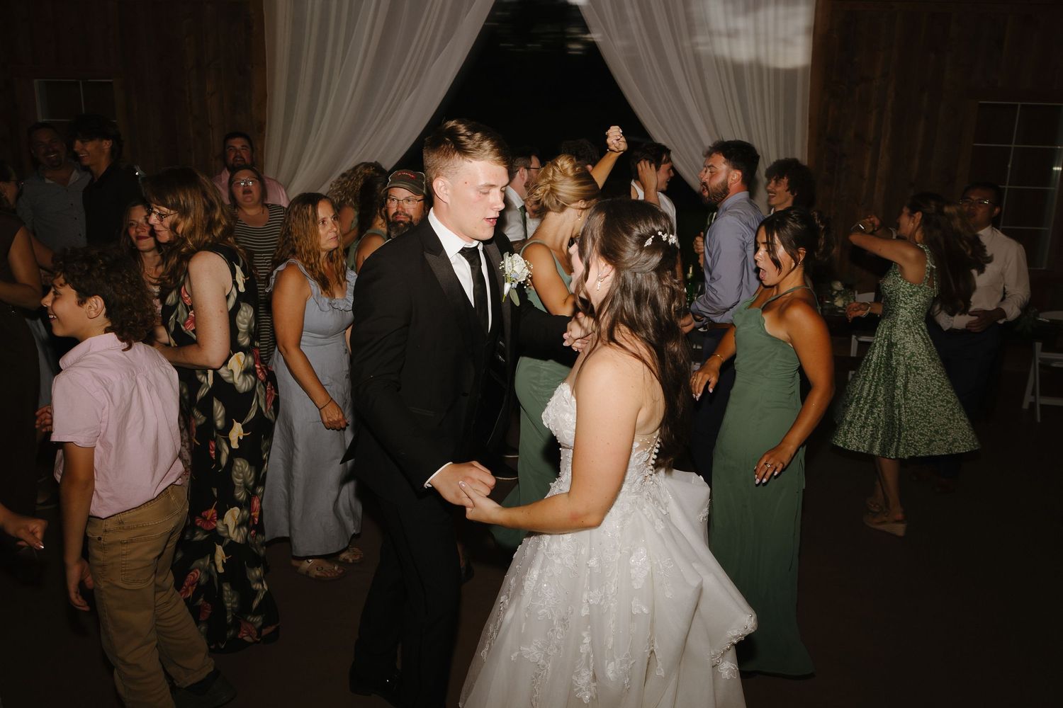 Bride and groom share first dance surrounded by wedding guests in rustic barn venue.