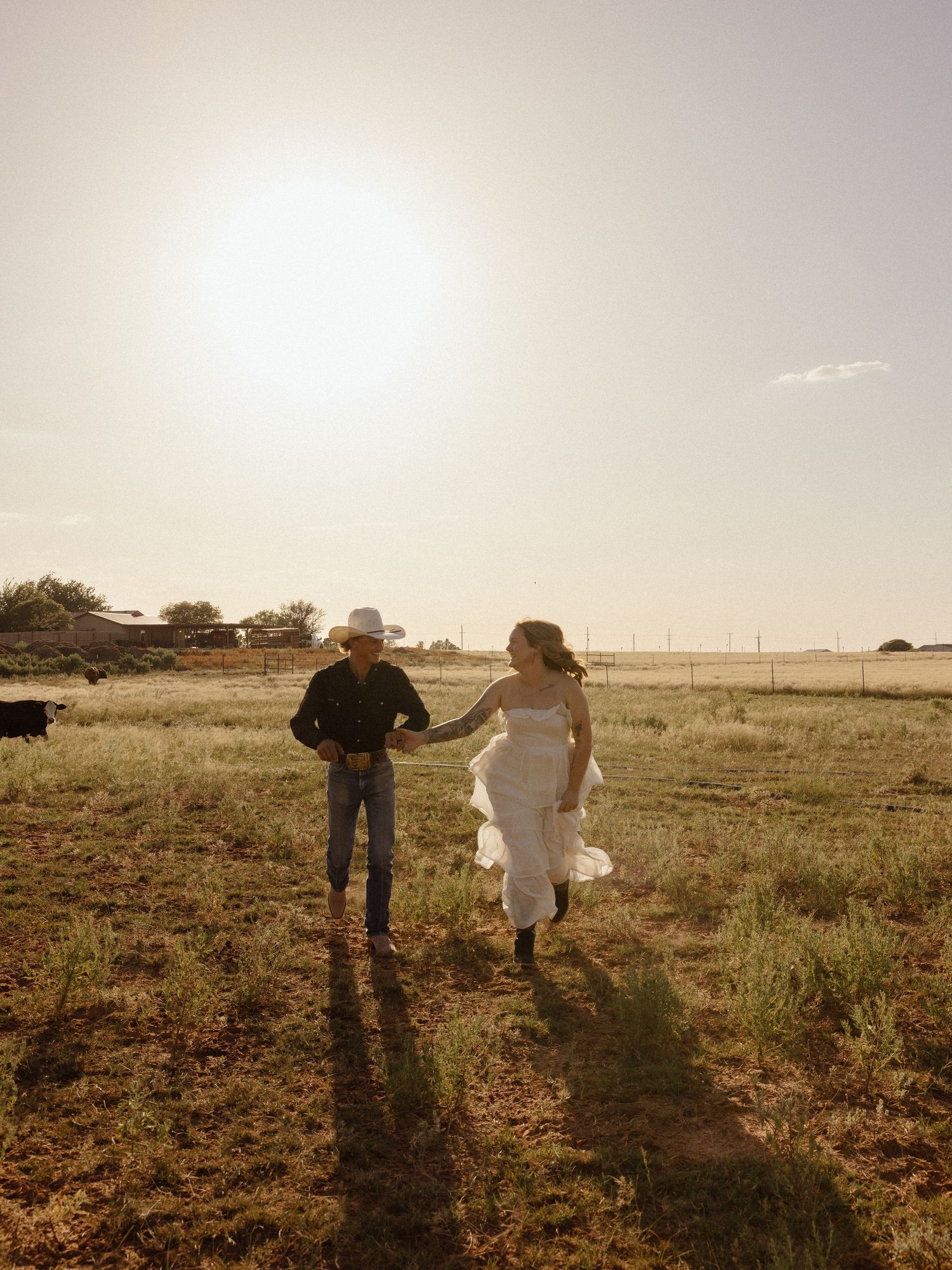 Couple running hand in hand through a bright sunlit open ranch field in western wedding attire.