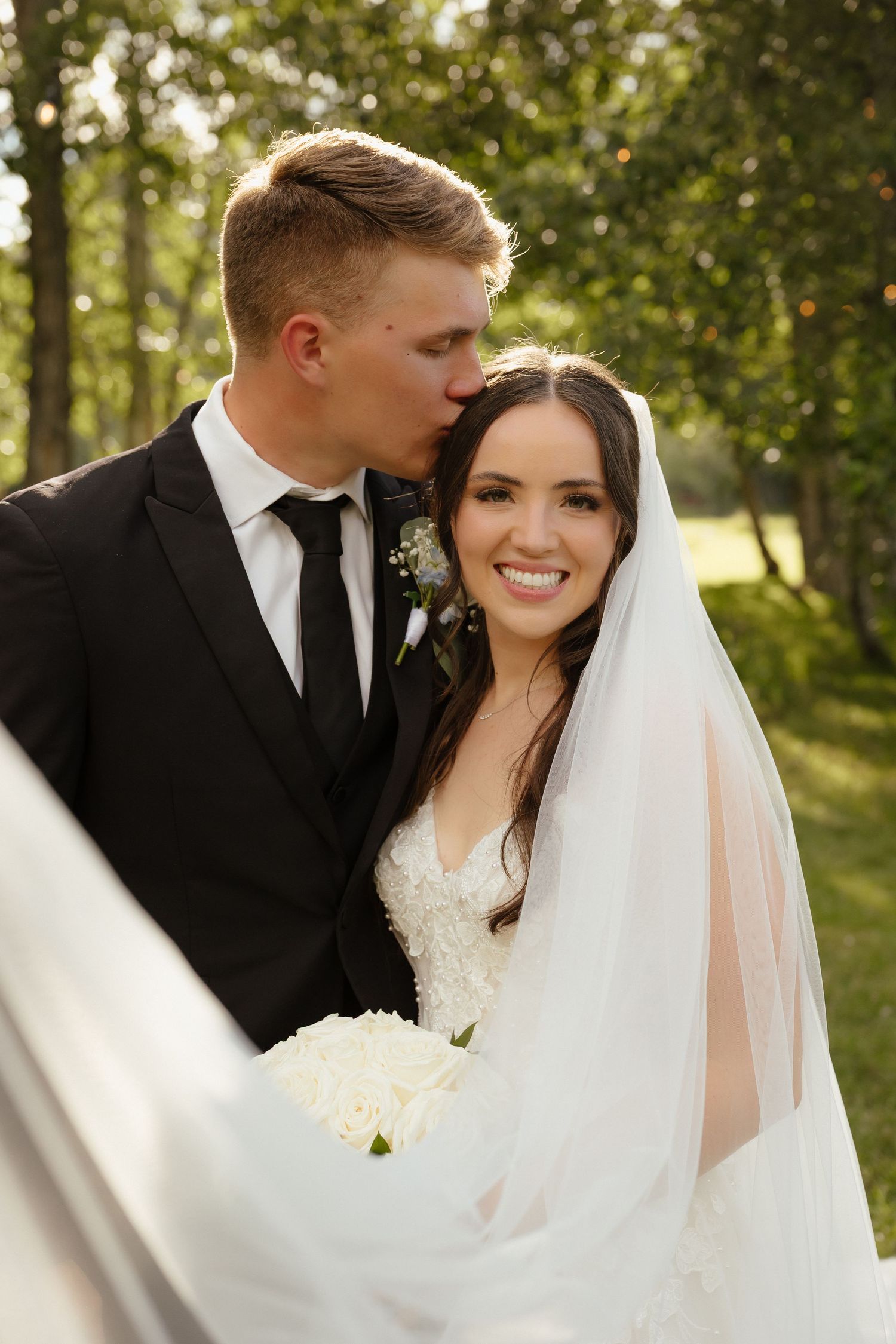 Groom in black suit kissing smiling bride on forehead while she holds white rose bouquet outdoors.