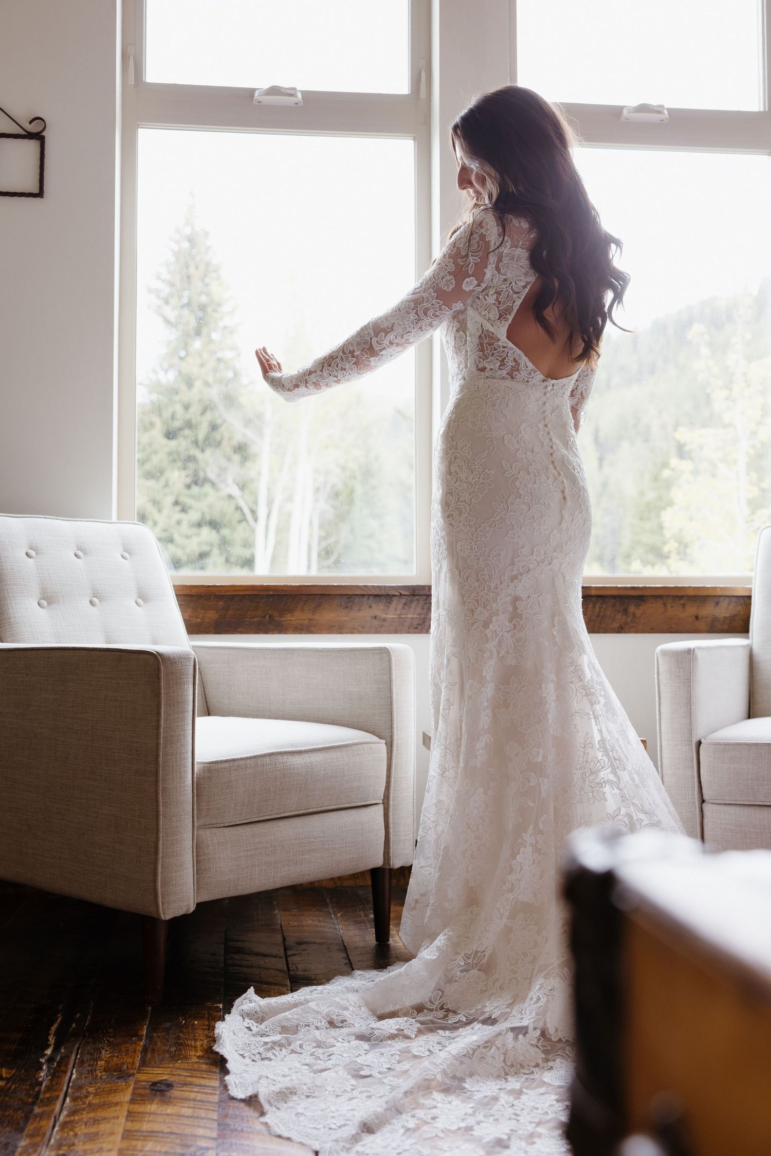 Bride in backless long-sleeve lace wedding gown with train standing by large windows in elegant venue.