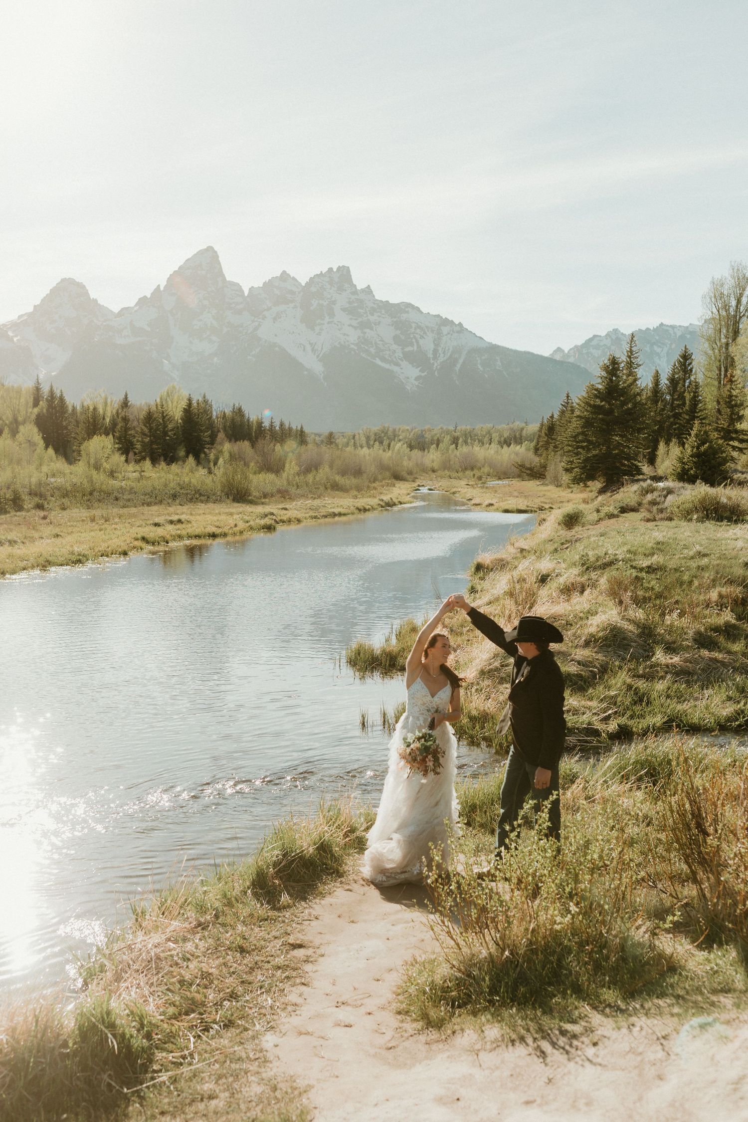 Western wedding couple dancing by scenic river with Grand Teton mountain backdrop at sunset.