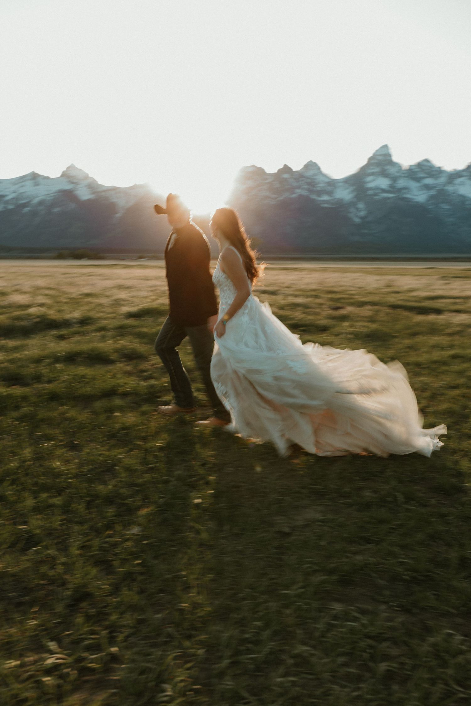 Silhouetted bride and groom walking in a meadow at golden hour with the Grand Teton mountains glowing behind them.