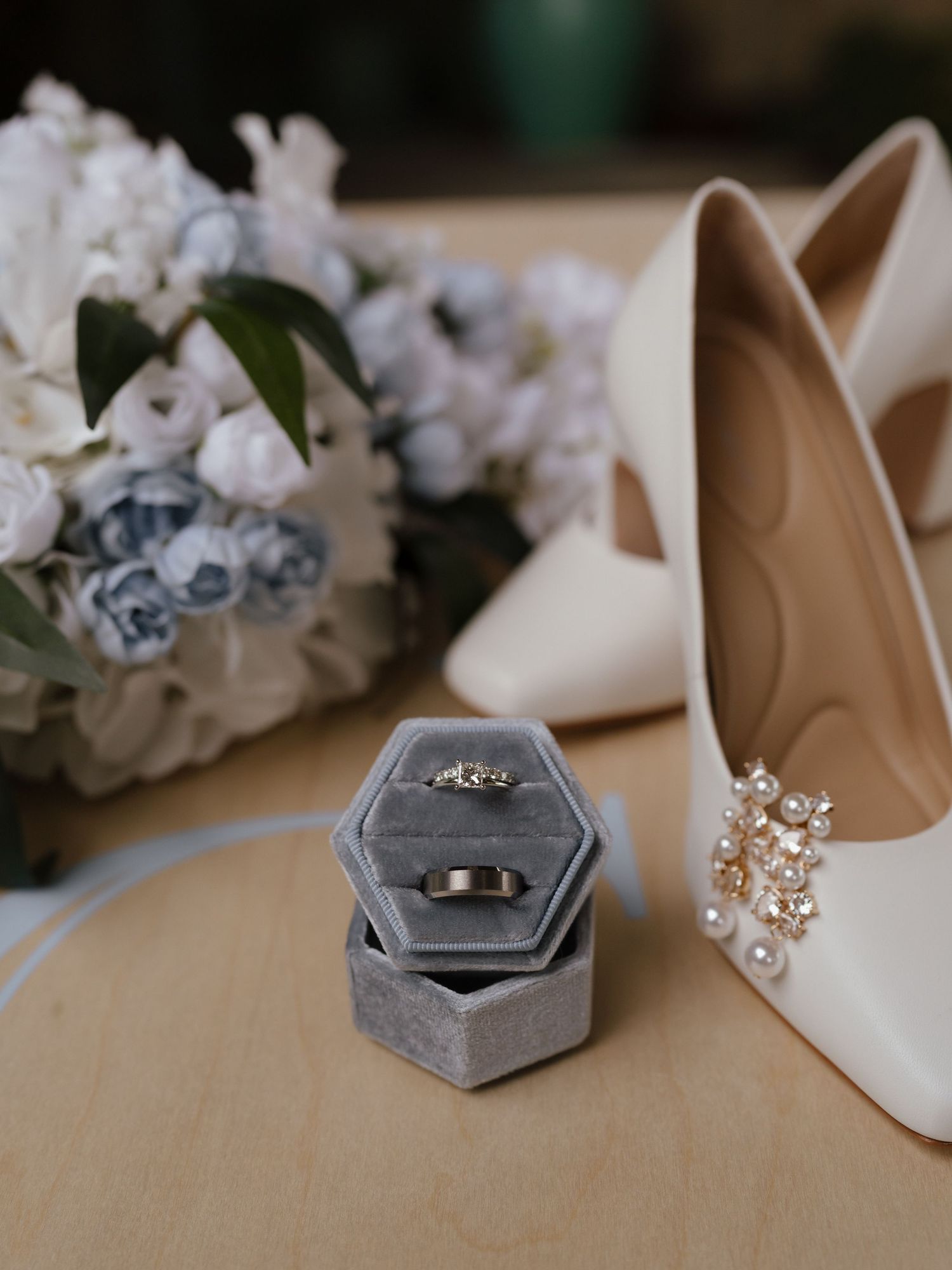 Gray velvet hexagon ring box with wedding bands beside pearl-embellished bridal heels and a white floral bouquet.