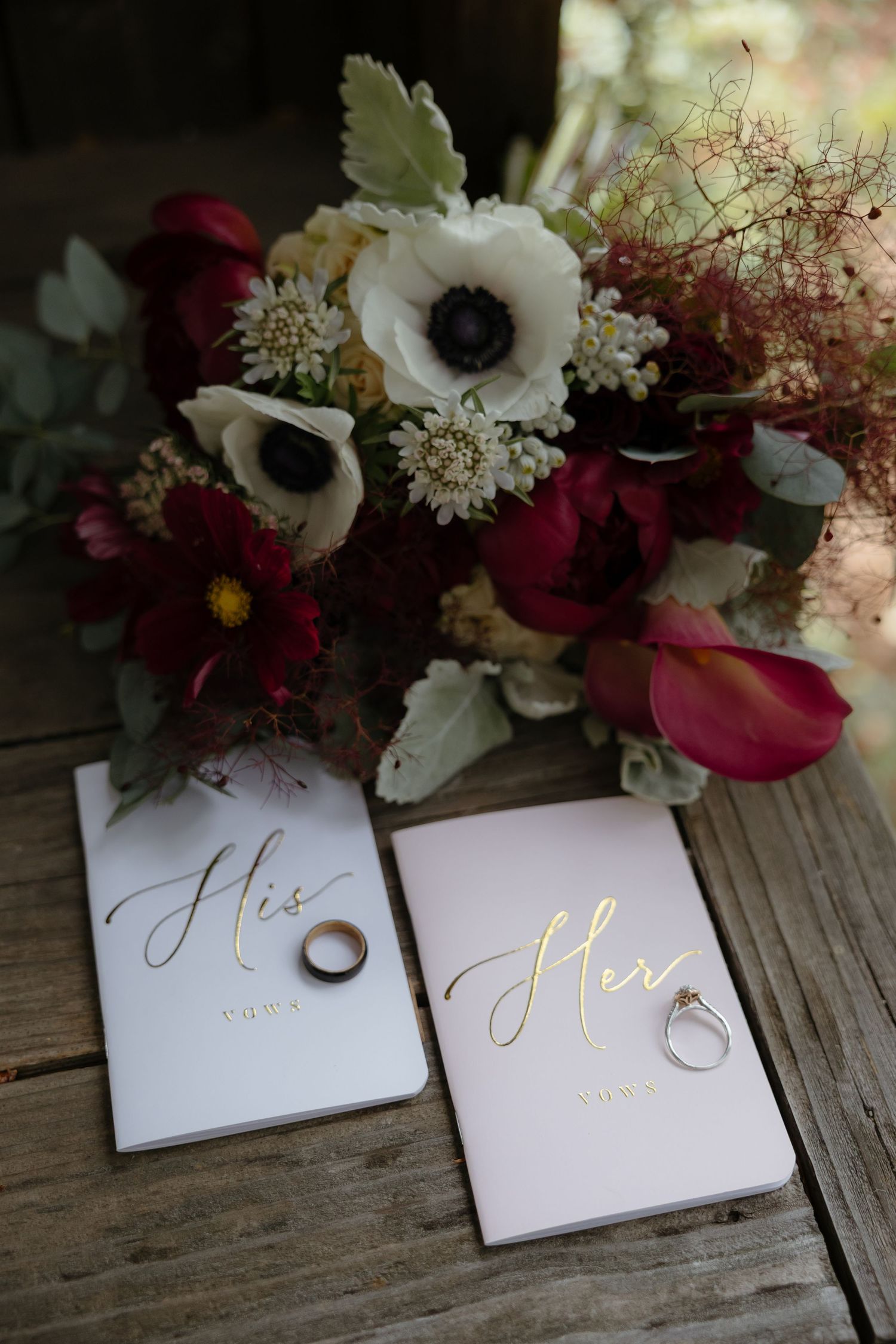 Wedding flat lay with burgundy and white floral bouquet, His and Hers vow books, and gold wedding rings on wood.