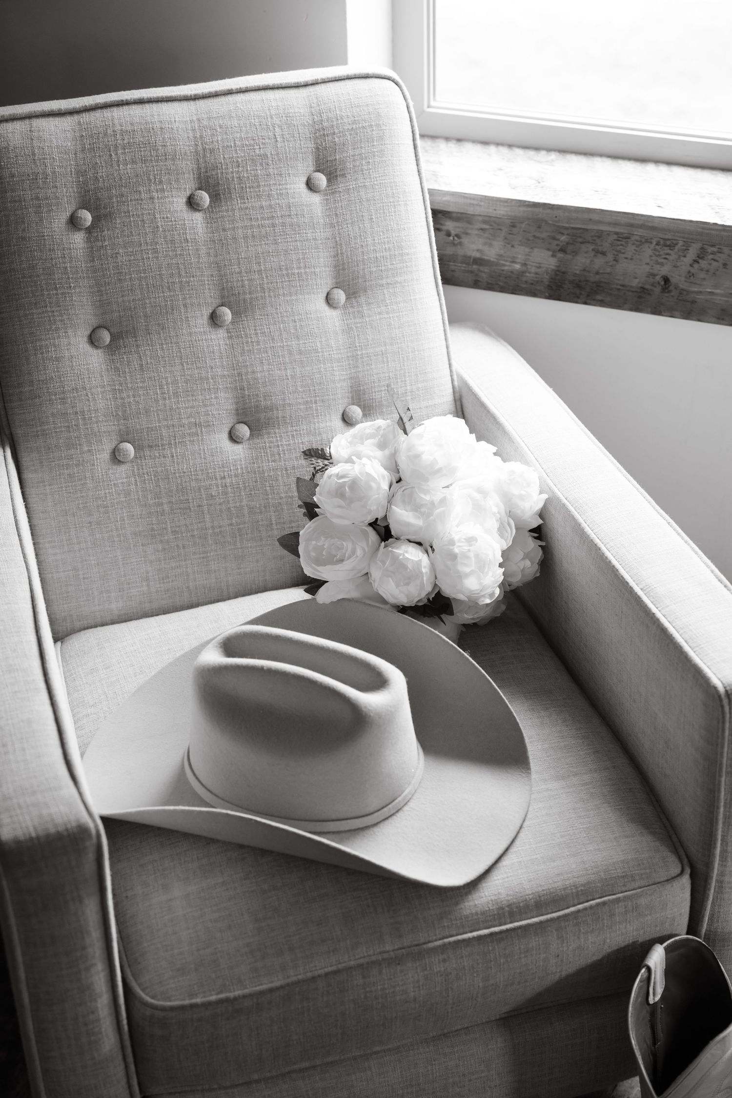 Black and white photo of a cowboy hat and white roses bouquet on a tufted chair by a window.