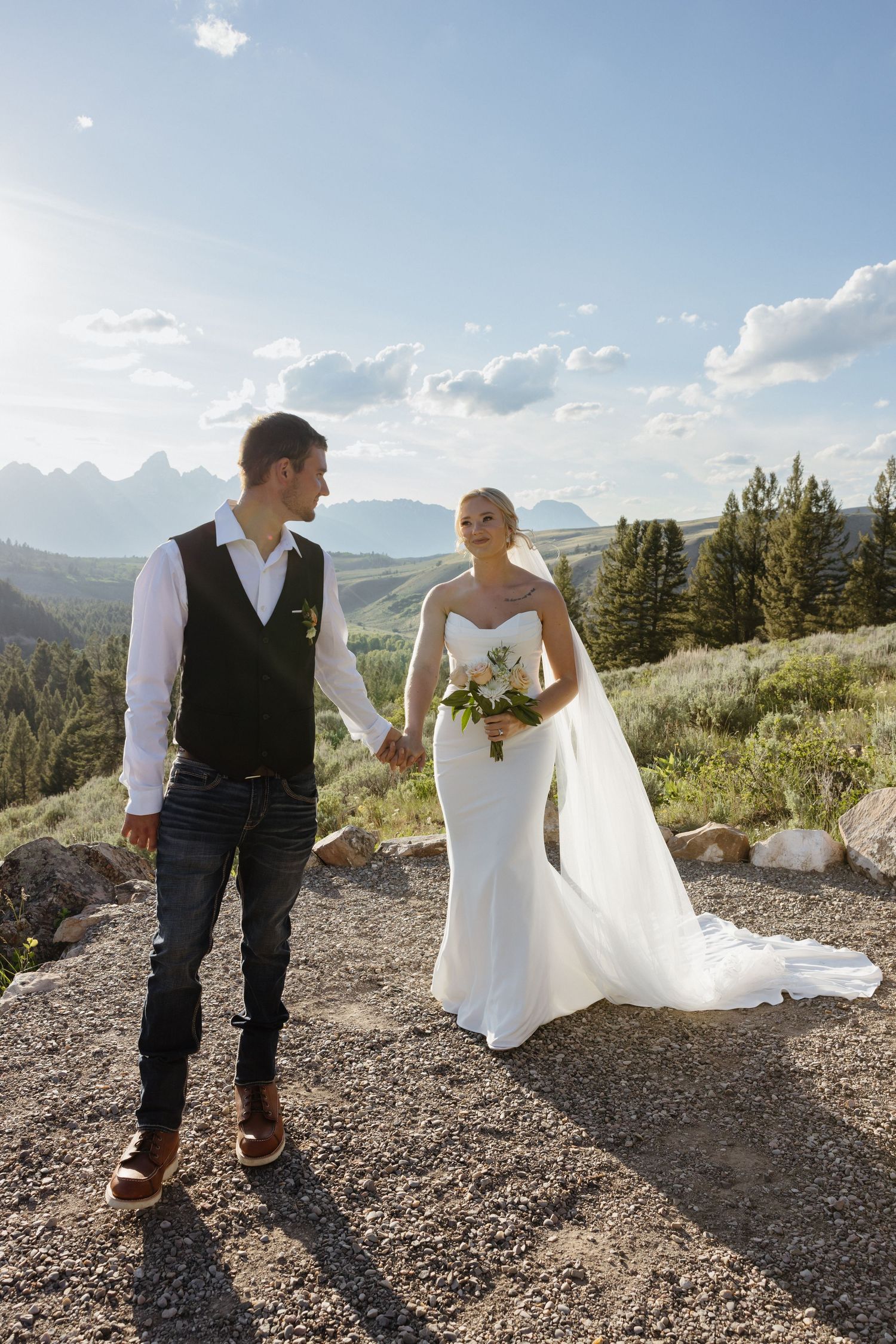 Bride and groom hold hands while walking on mountain overlook with Grand Teton peaks under blue sky.