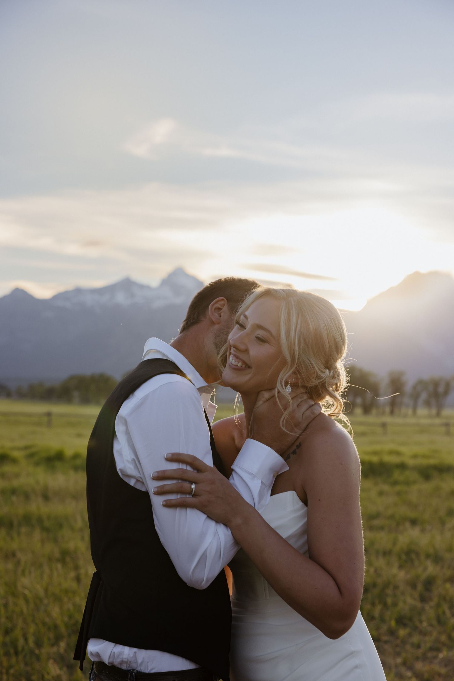 Joyful bride laughs as groom kisses her cheek during golden hour with mountain landscape glowing behind them.