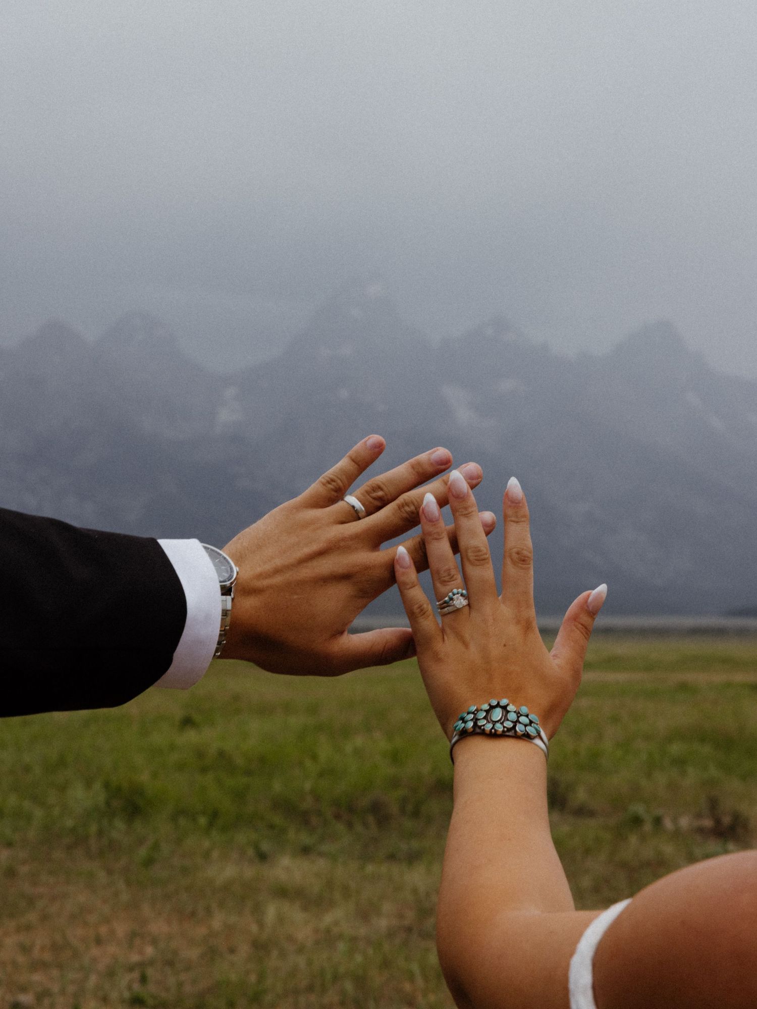 Couple displaying wedding rings with misty mountain backdrop, bride wearing turquoise bracelet and elegant engagement ring.