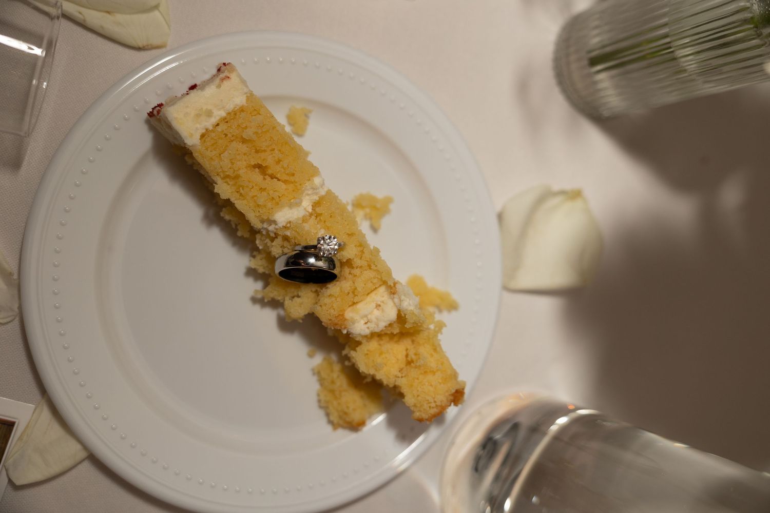 Slice of white wedding cake with small ring decoration on white plate at reception.