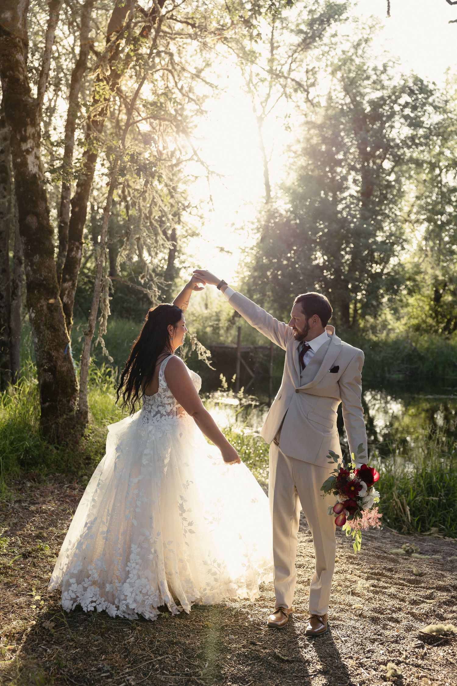 Groom spinning bride in elegant lace wedding dress during golden sunset portraits near a tranquil forest pond.