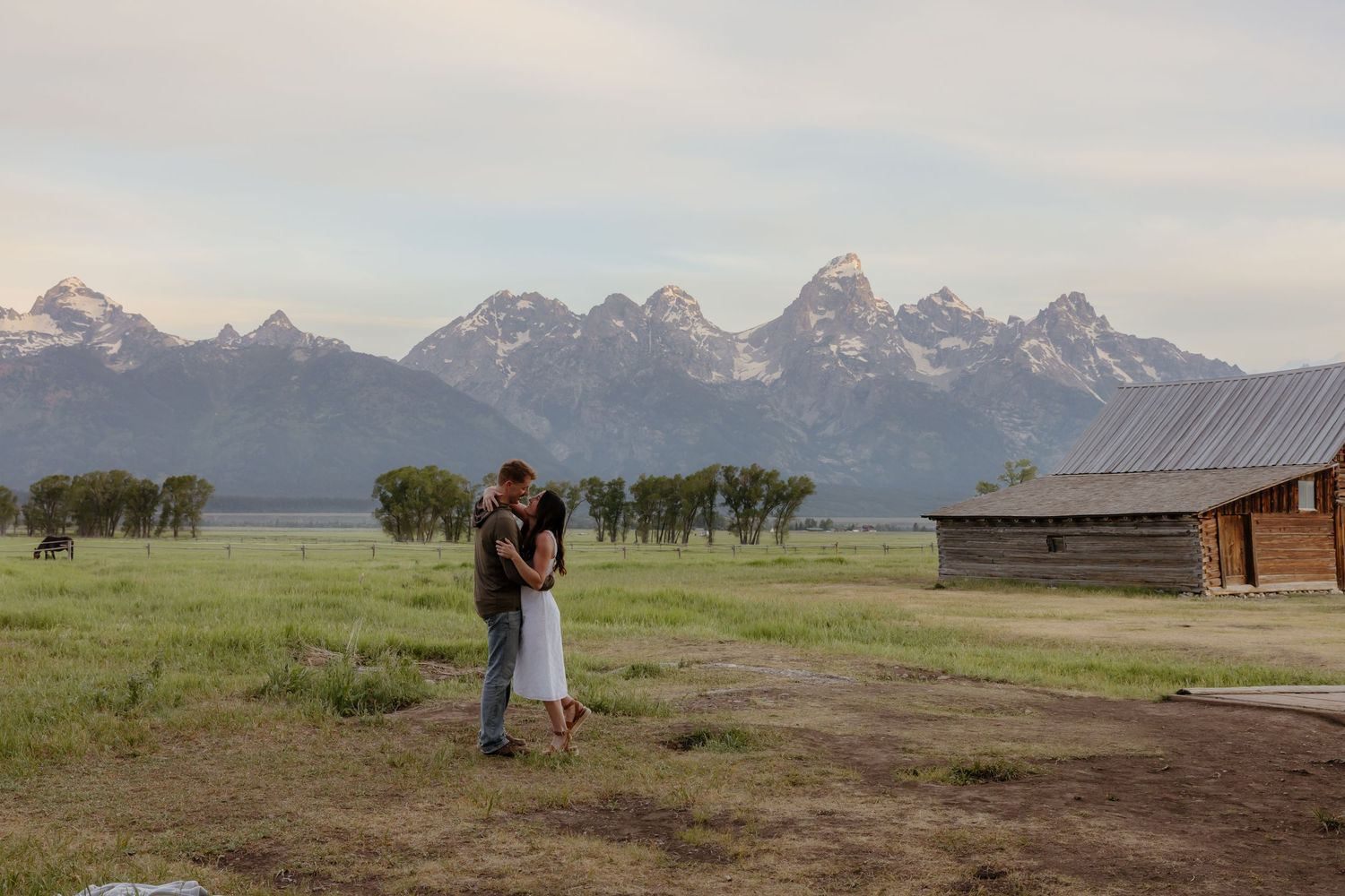 Couple embracing near a rustic barn with the iconic Grand Teton mountain range stretching across the horizon.