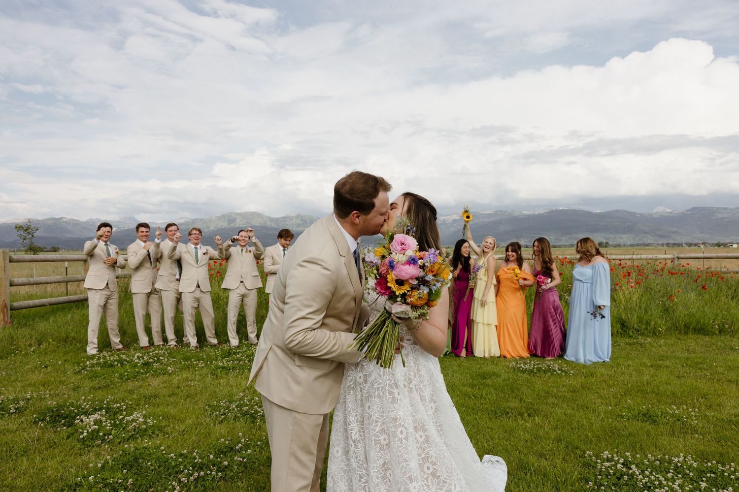 Newlyweds share a kiss while wedding party in colorful dresses and tan suits celebrate in mountain field.