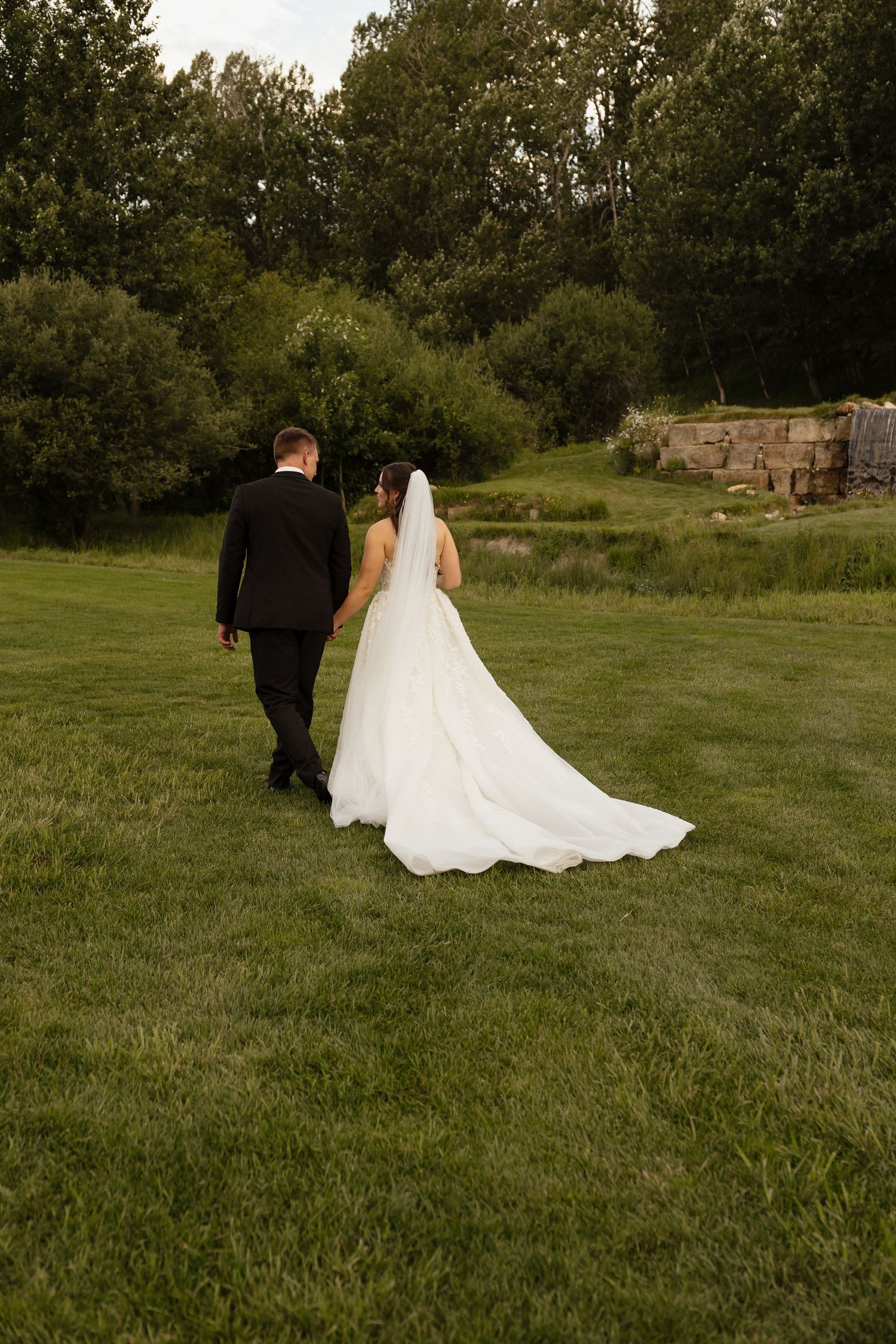 Bride and groom walking hand in hand across green lawn with long white dress train trailing behind.