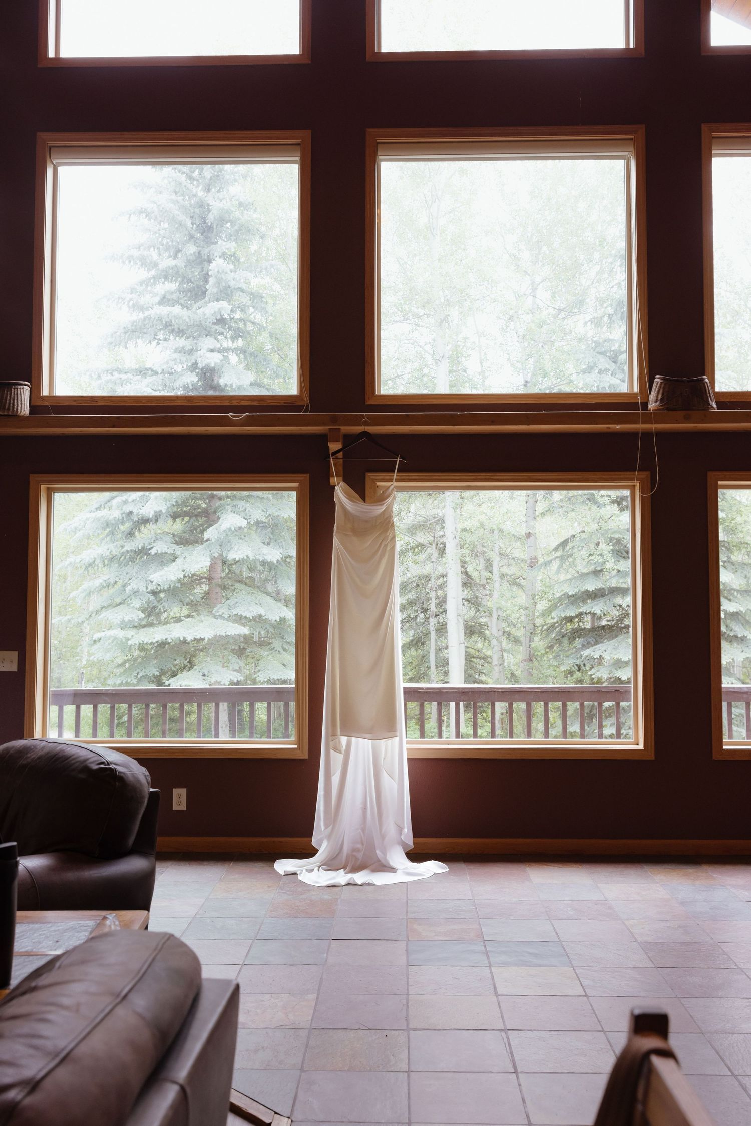 Elegant white wedding dress hangs in front of large cabin windows overlooking lush green pine trees outside.
