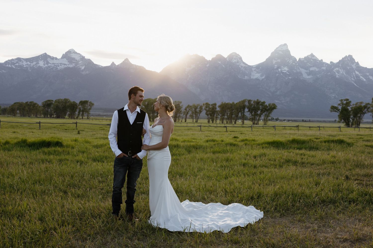 Bride and groom standing in green meadow at golden sunset with snow-capped Grand Teton peaks.