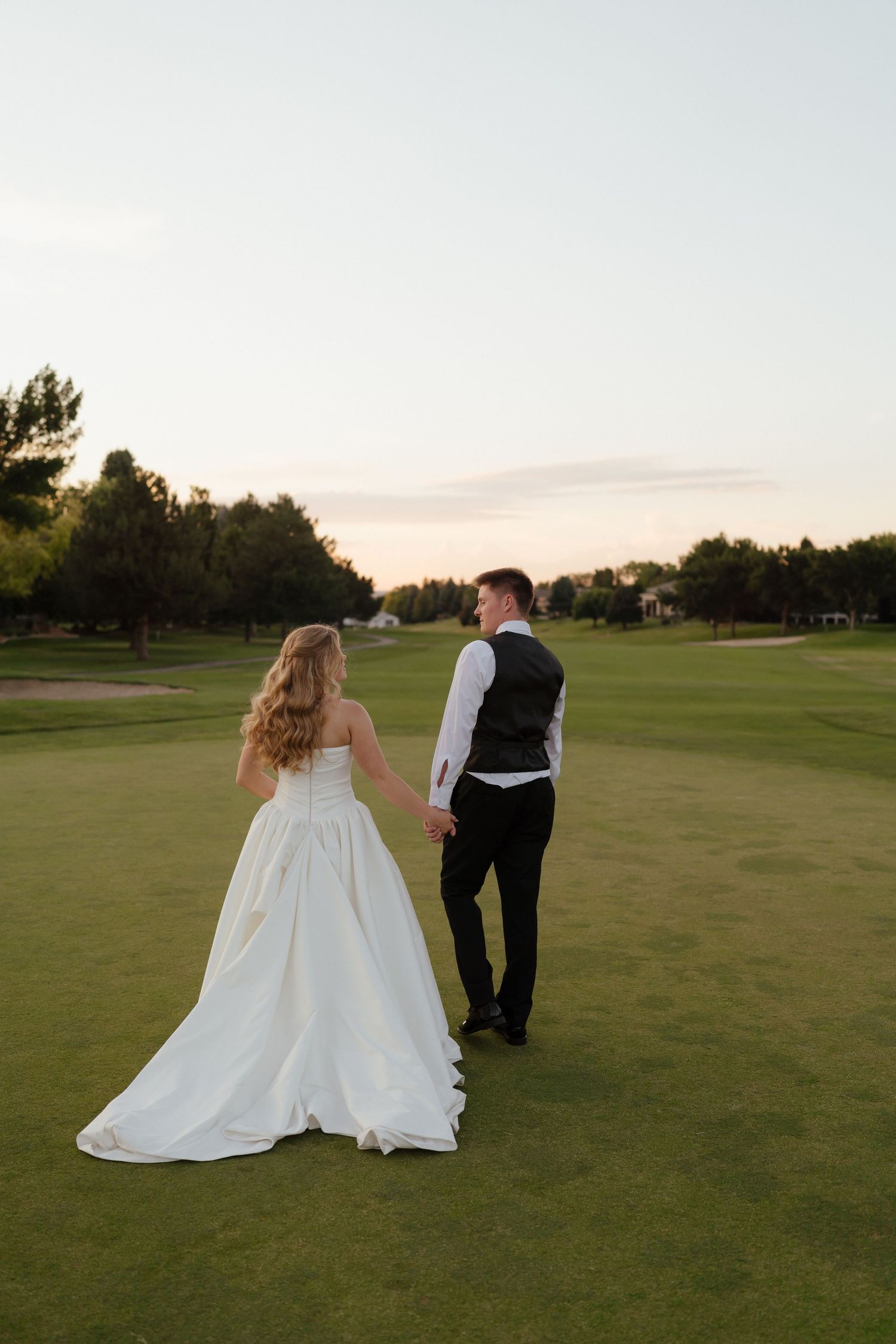 Bride and groom holding hands walking on lush green golf course at sunset in wedding attire.