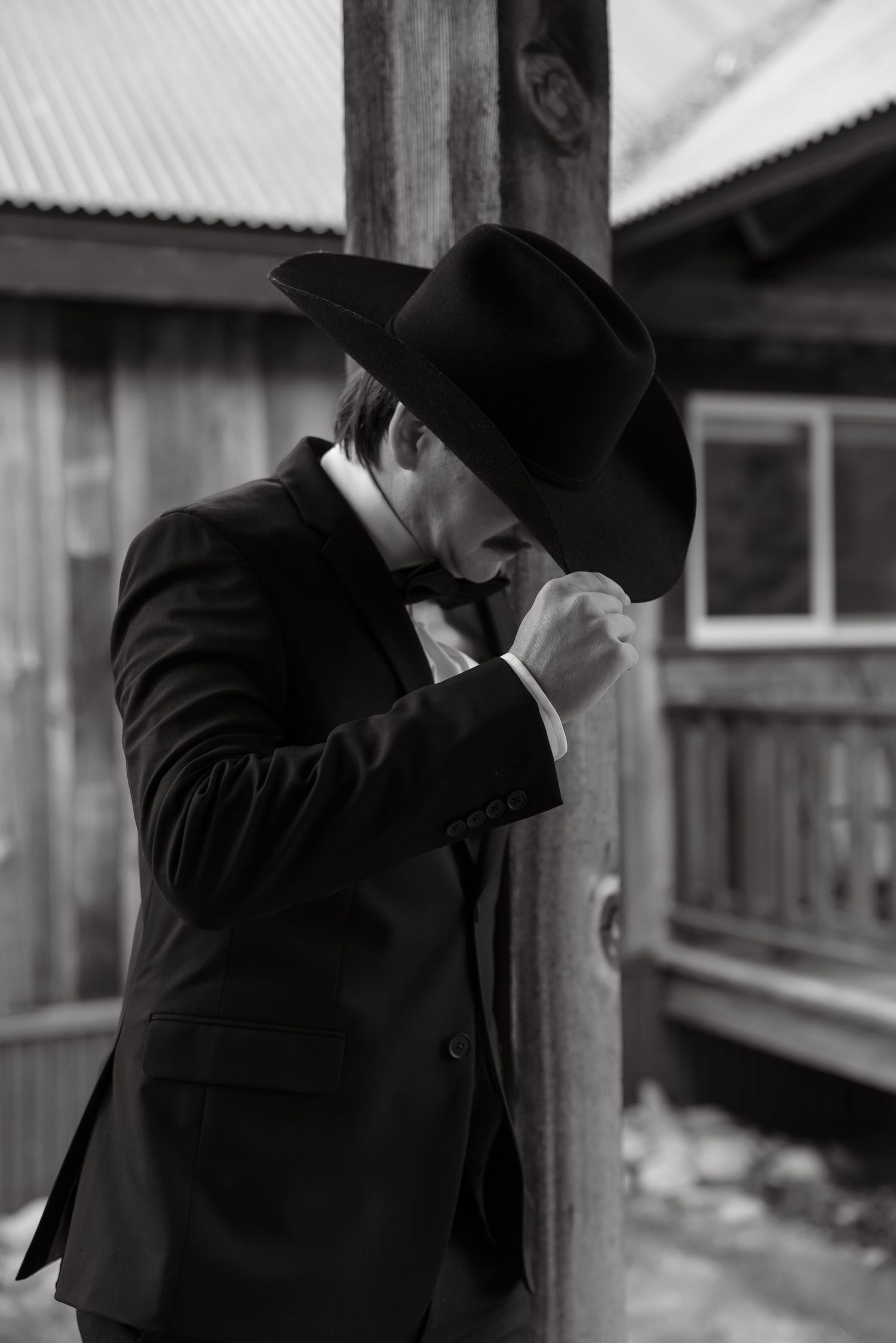 Black and white portrait of groom in black suit and cowboy hat leaning against a wooden post.