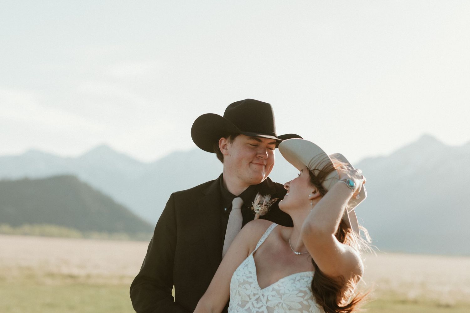 Bride and groom sharing a romantic moment in a mountain meadow, groom wearing a black cowboy hat and suit.