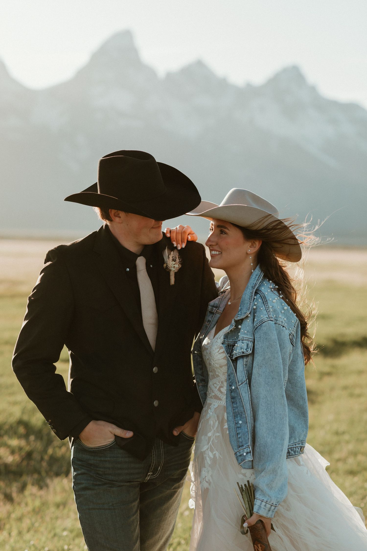 Bride in denim jacket and white cowboy hat smiles at groom in black hat with Grand Teton mountains behind them.