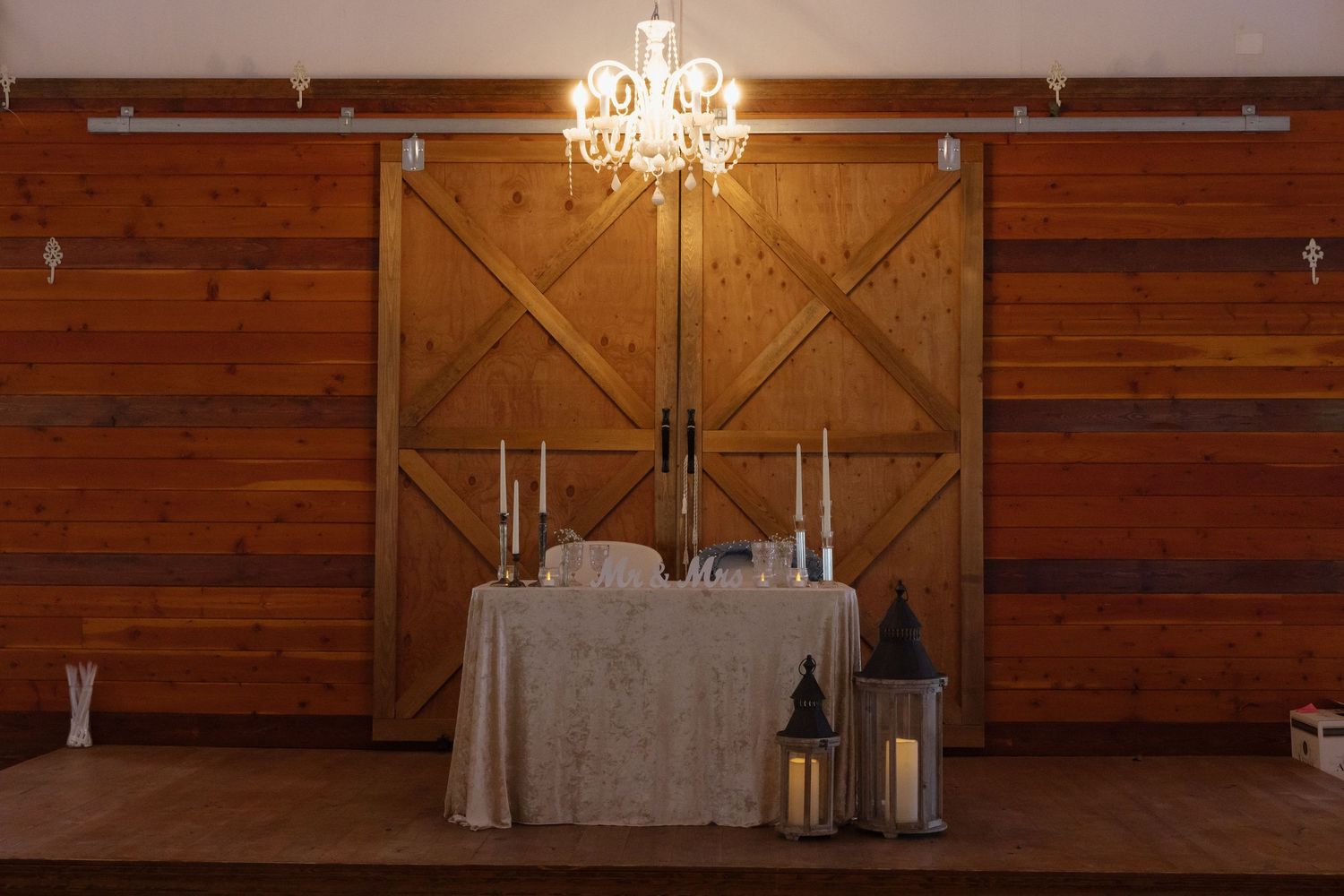 Rustic barn wedding sweetheart table with candlesticks and lanterns in front of wooden sliding barn doors with chandelier.
