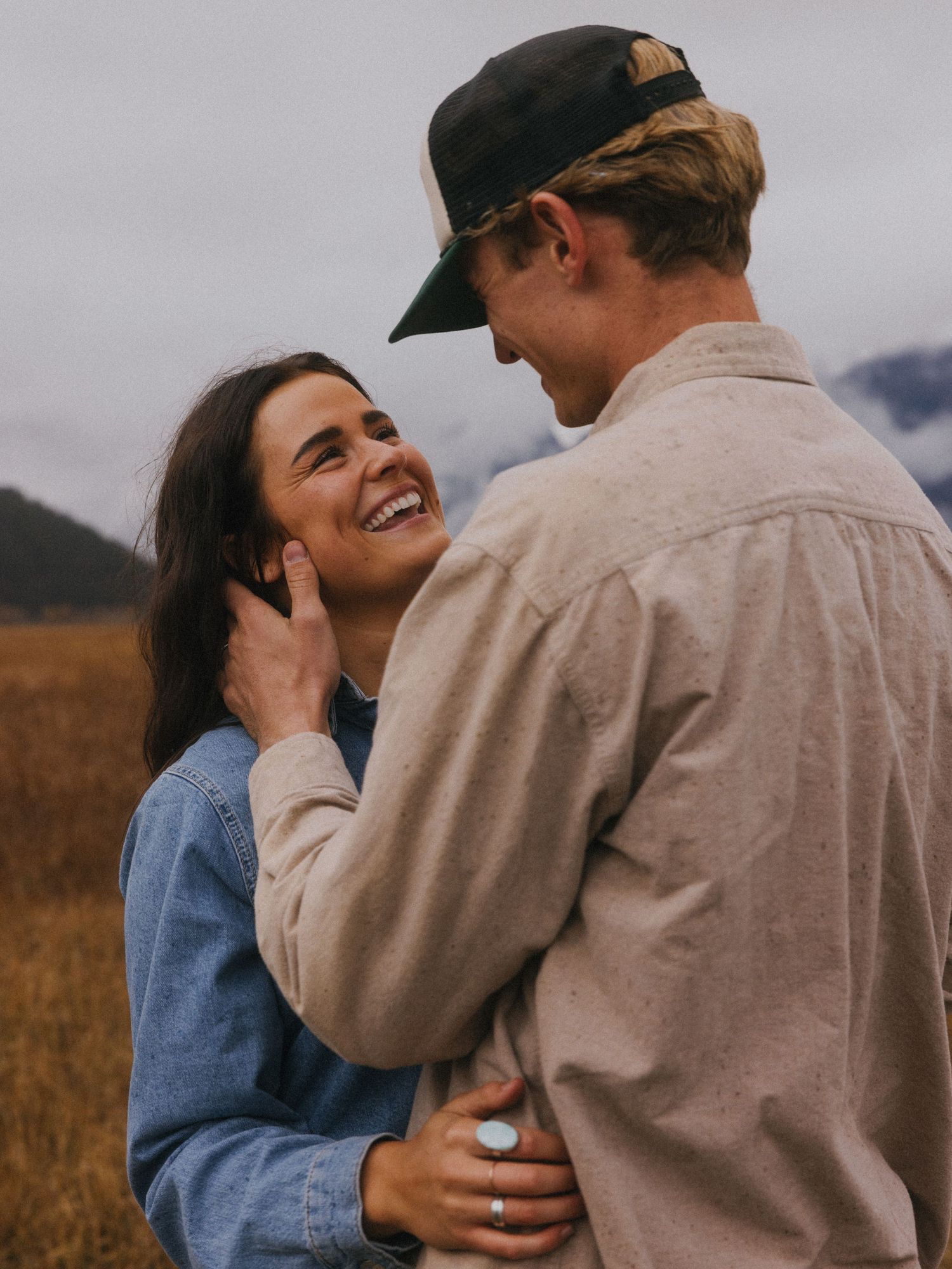 Smiling woman gazes at her partner while embracing in a field with mountains in the background.