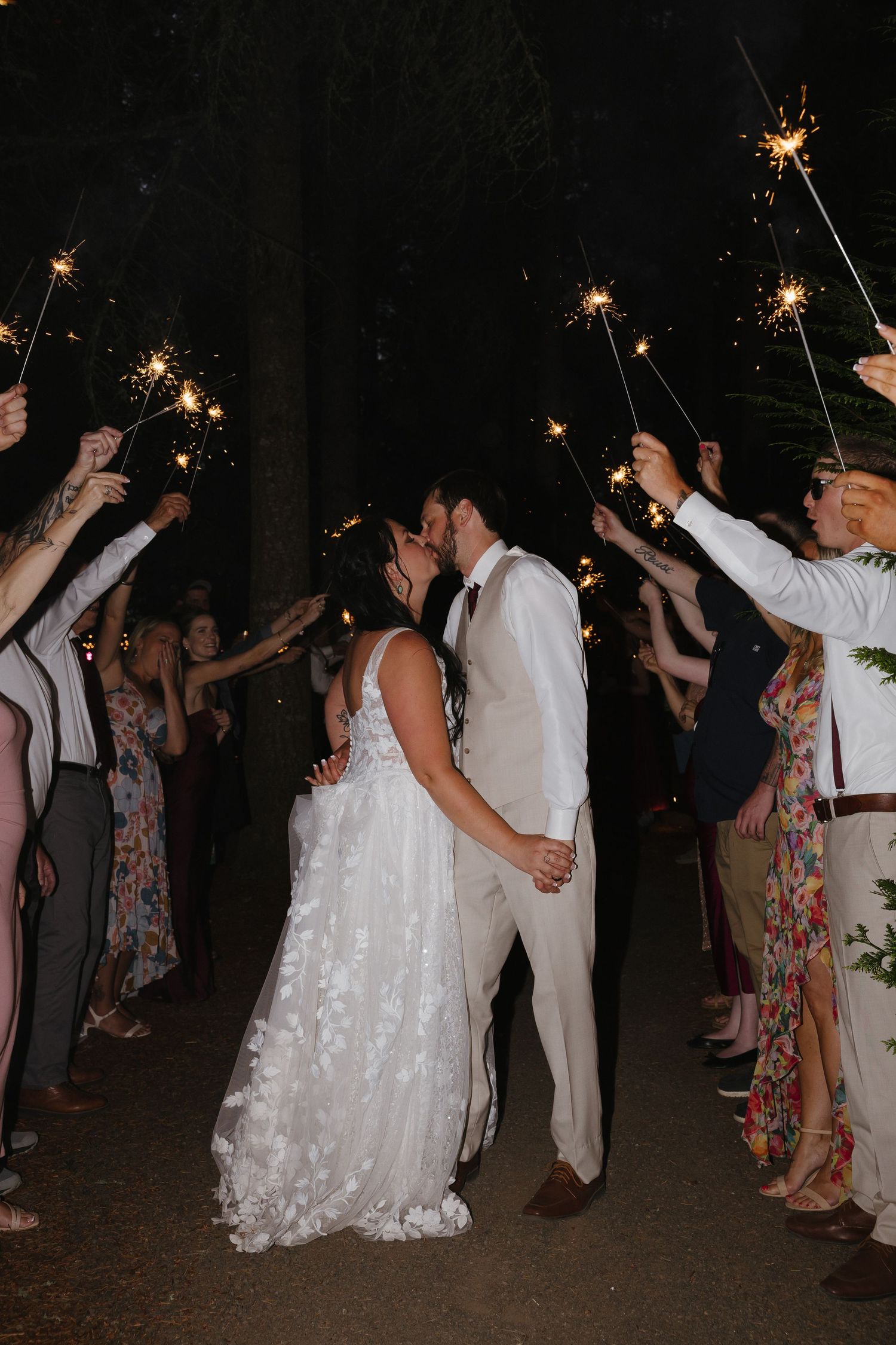 Bride and groom kiss during sparkler send-off exit lined by cheering guests at nighttime outdoor forest wedding.