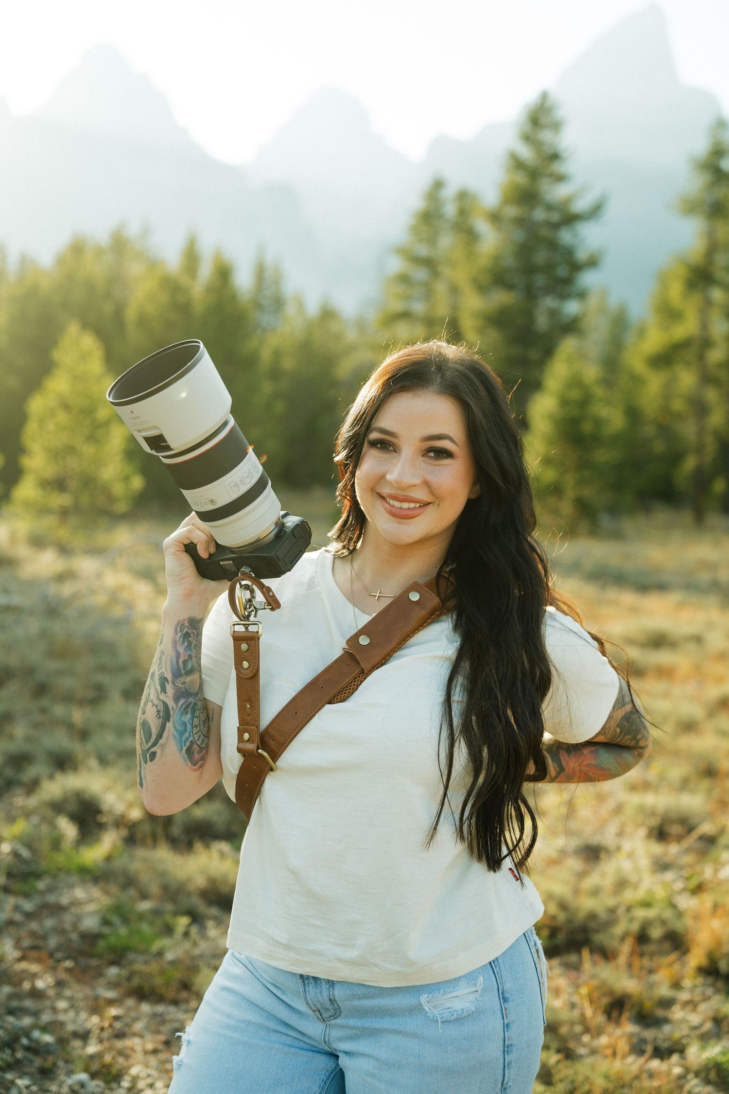 Tattooed female photographer smiles holding professional Canon telephoto camera with Grand Teton mountains glowing behind her.