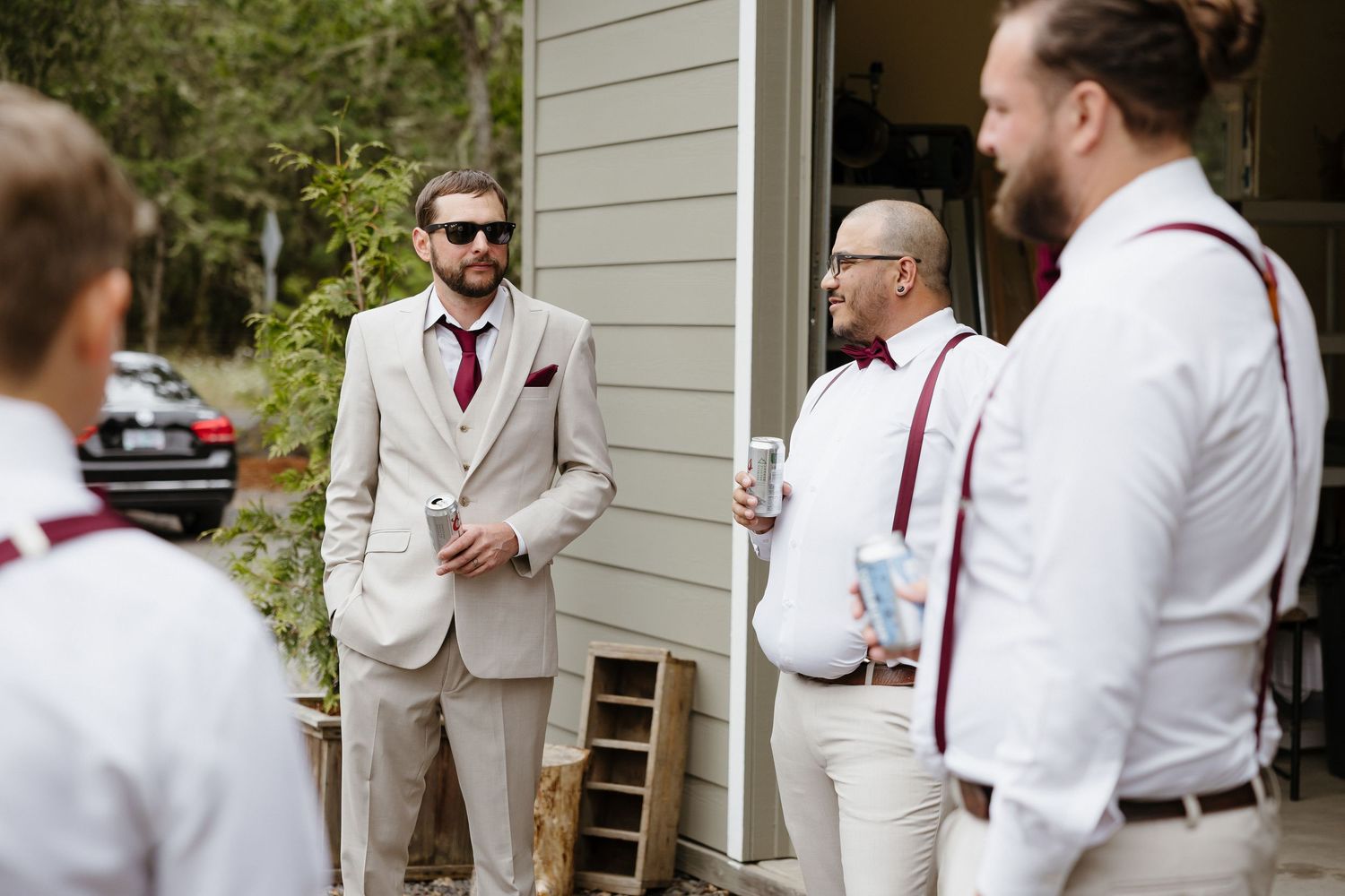 Groom in tan suit with burgundy tie and groomsmen in suspenders gathering outside before wedding.