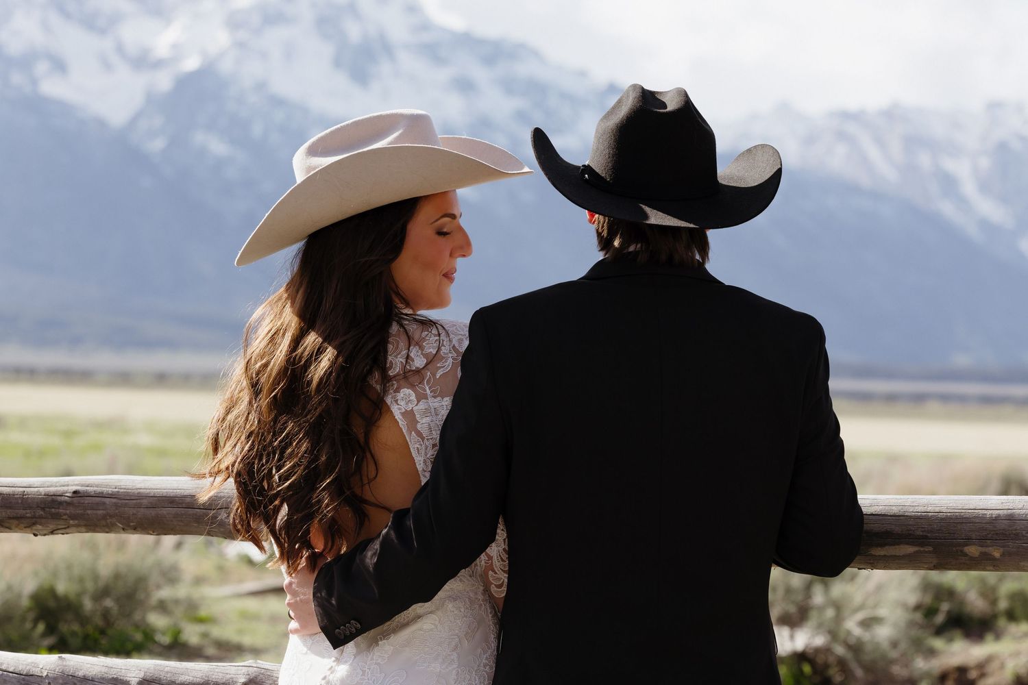 Bride and groom in cowboy hats stand by a rustic fence with mountain views at their western-themed wedding.
